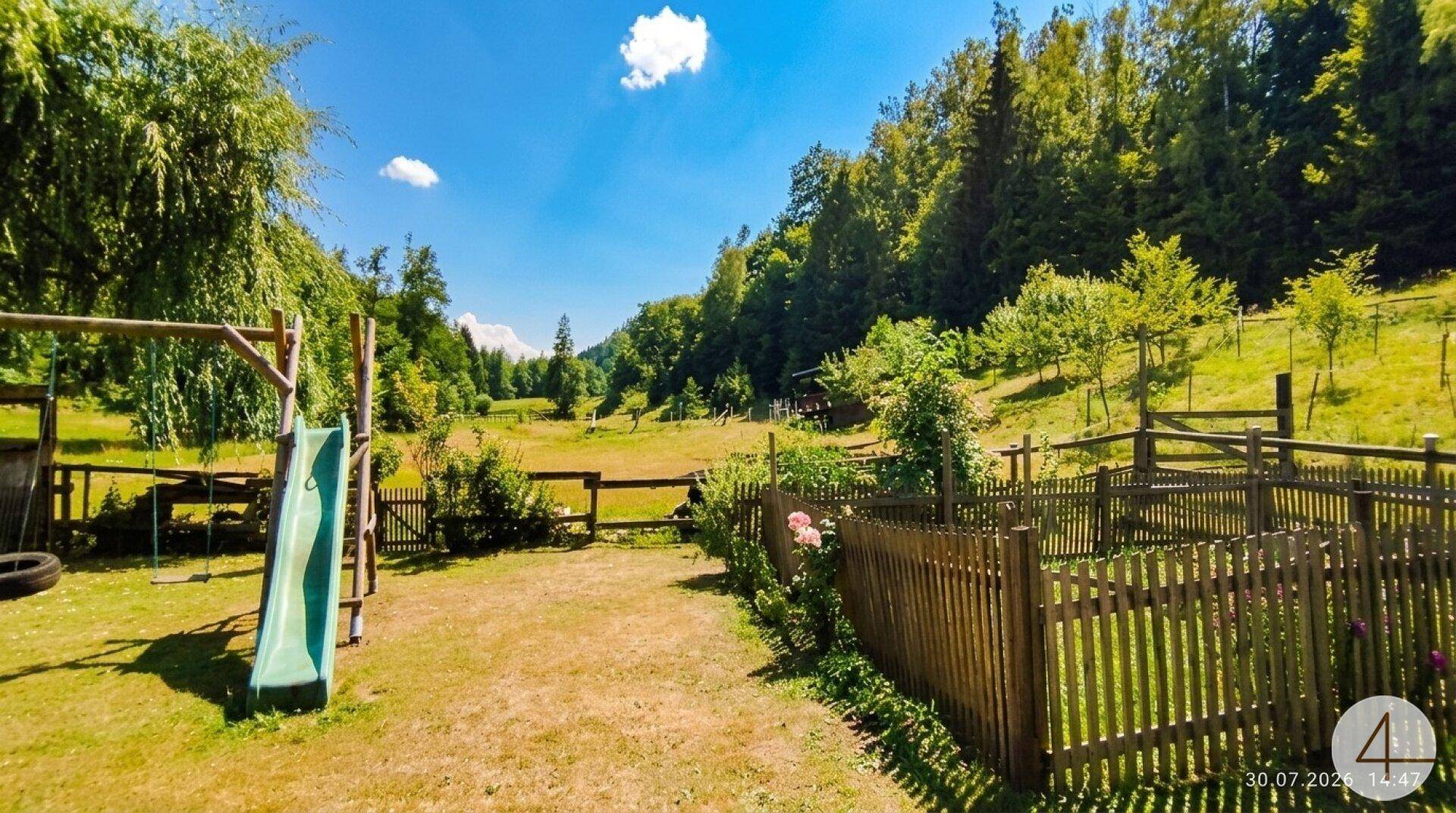 Großer Spielplatz mit Rutsche und Schaukeln, umgeben von einem Holzzaun und grüner Landschaft.