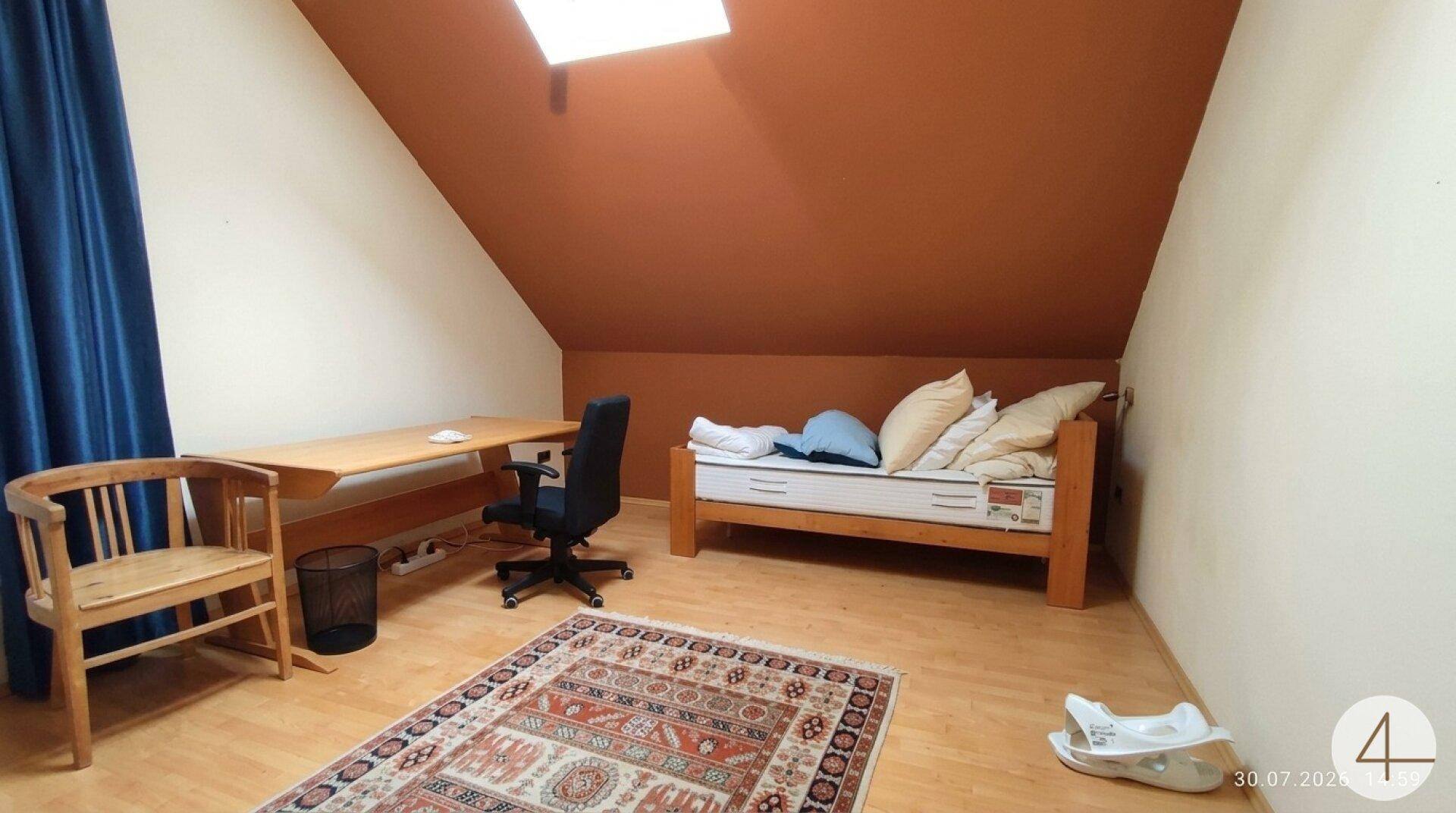 Gemütliches Schlafzimmer im Dachgeschoss mit Holzbett, Schreibtisch und Teppich, beleuchtet durch ein Dachfenster.