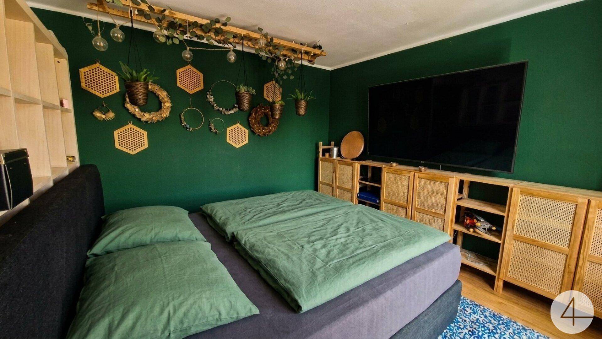 Gemütliches Schlafzimmer mit dunkelgrünen Wänden, großem Fernseher und Holzelementen.