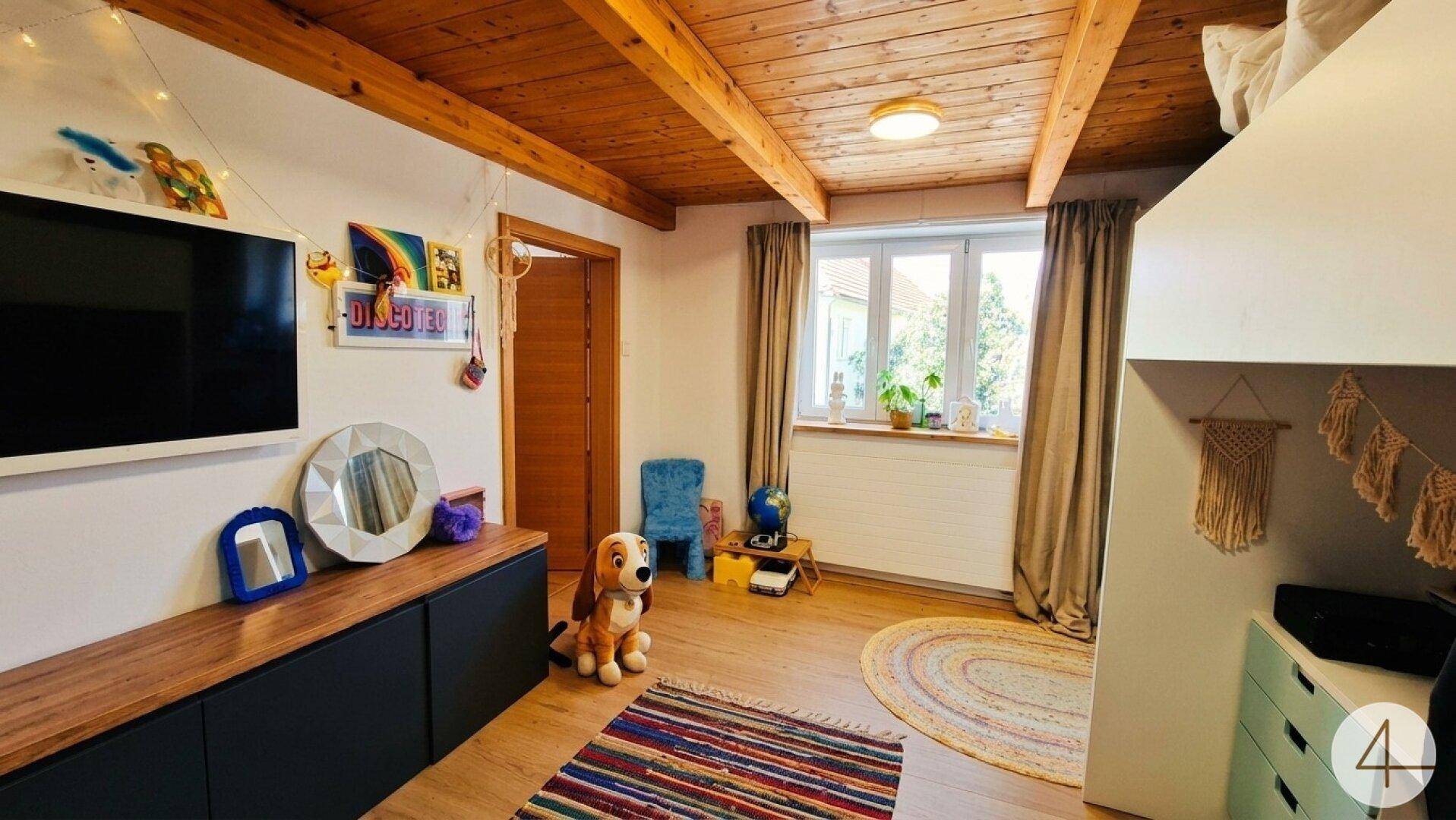 Gemütliches Kinderzimmer mit Holzbalkendecke, Fenster und bunter Dekoration.
