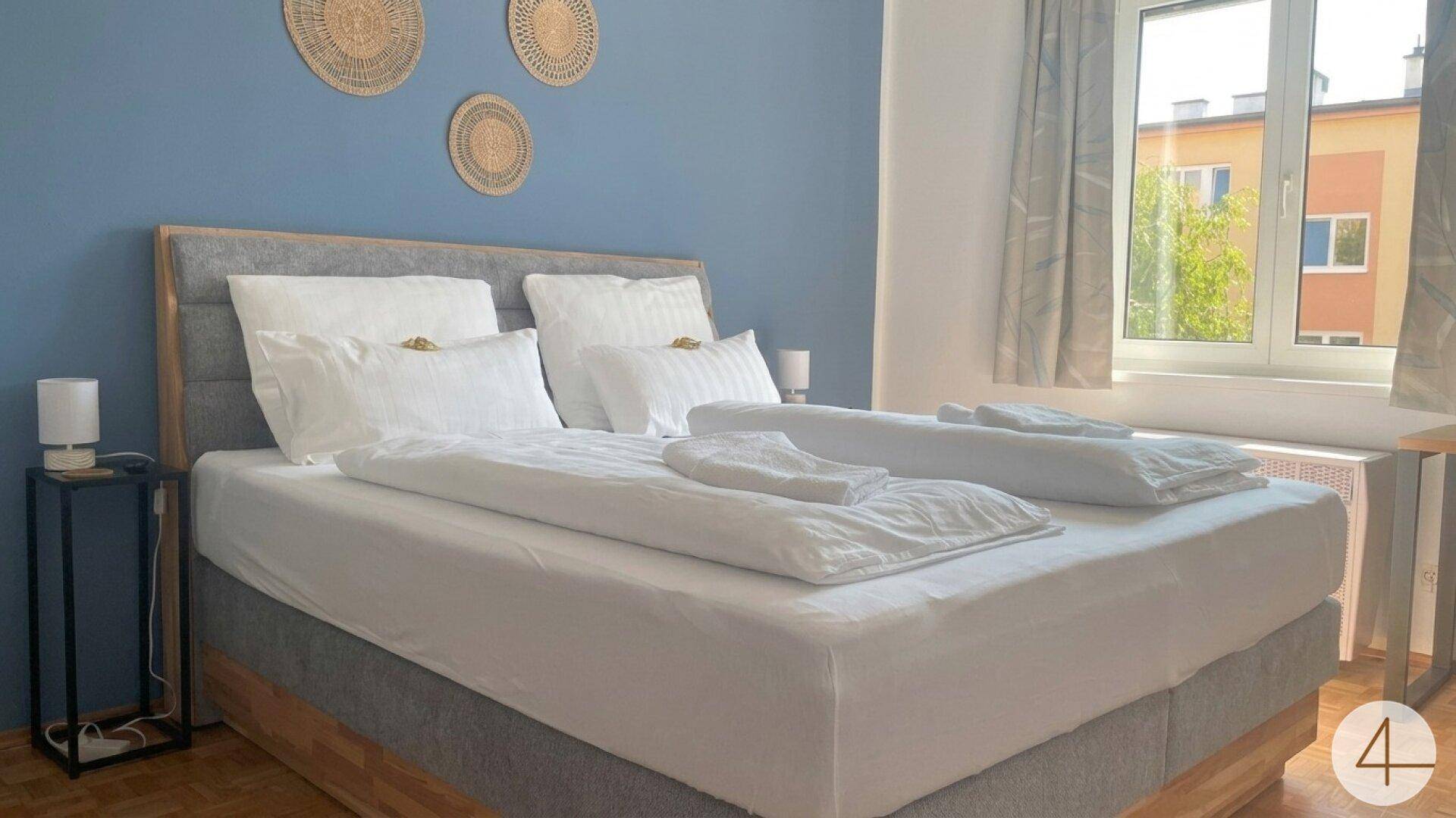 Schlafzimmer mit Doppelbett, blauer Akzentwand und Fenster, das viel Tageslicht hereinlässt.