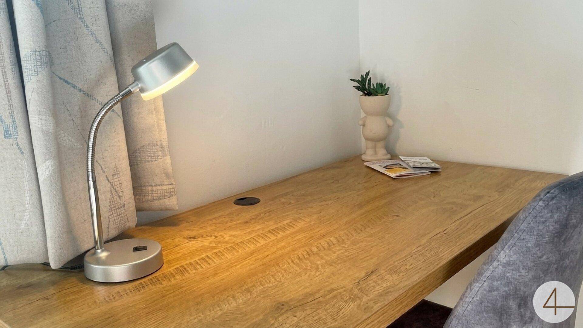 Heller Arbeitsbereich mit Schreibtisch, flexibler Lampe und kleiner Dekoration am Fenster.