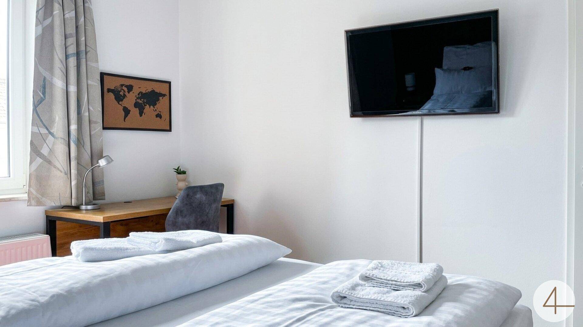 Schlafzimmer mit bequemem Bett, wandmontiertem Fernseher und einem kleinen Arbeitsbereich.