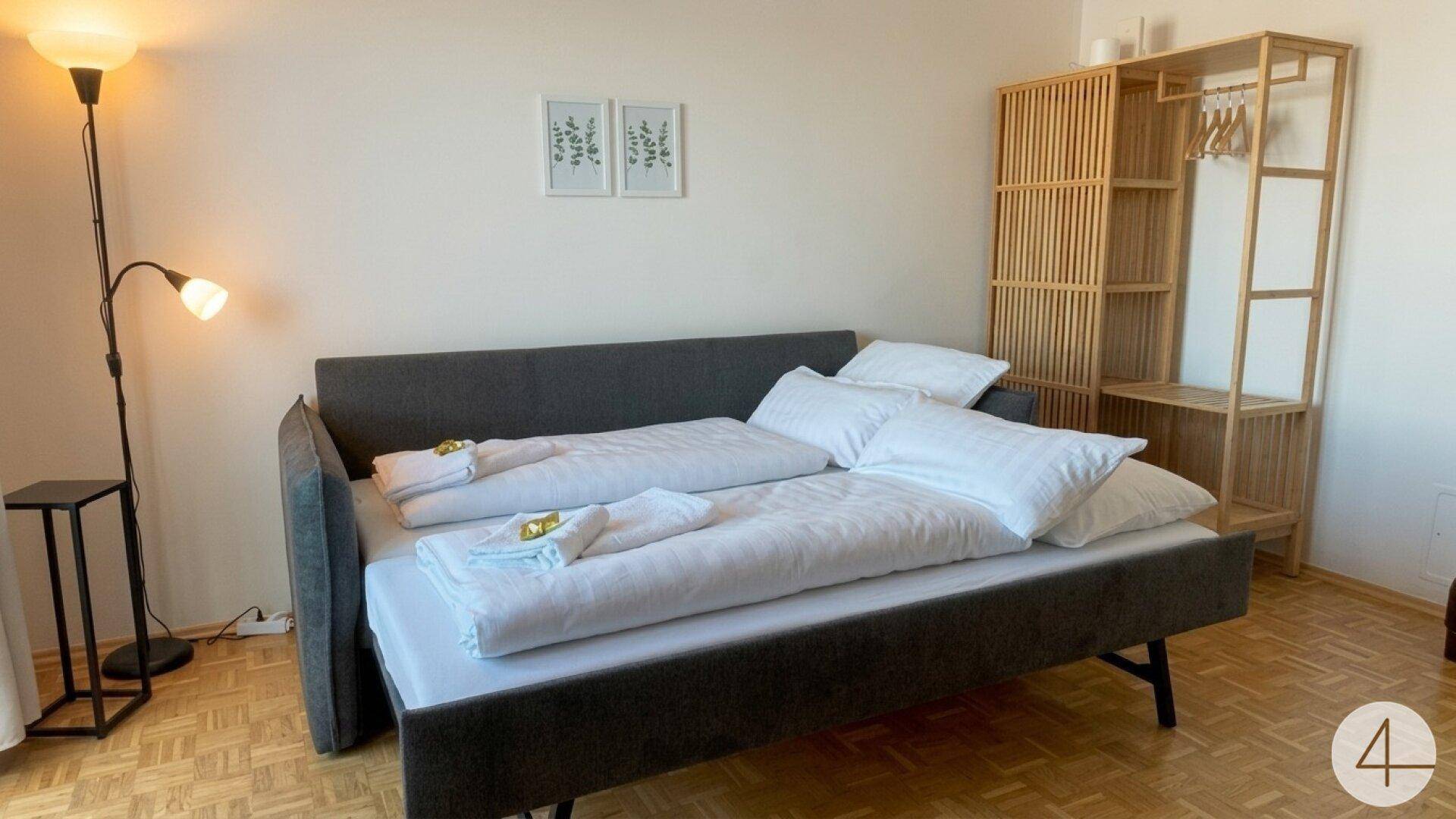 Schlafsofa im Wohnzimmer, als Bett hergerichtet, mit Stehlampe und offenem Holzregal.