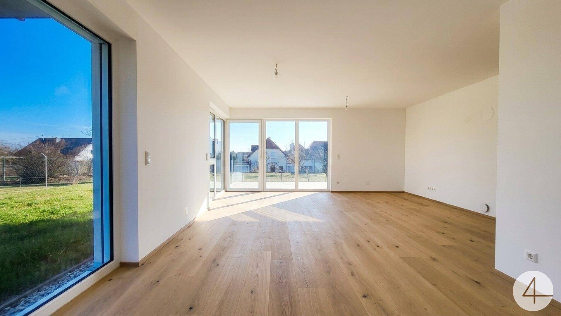 Heller, leerer Wohnraum mit Holzboden und großen Fenstern mit Blick ins Grüne.