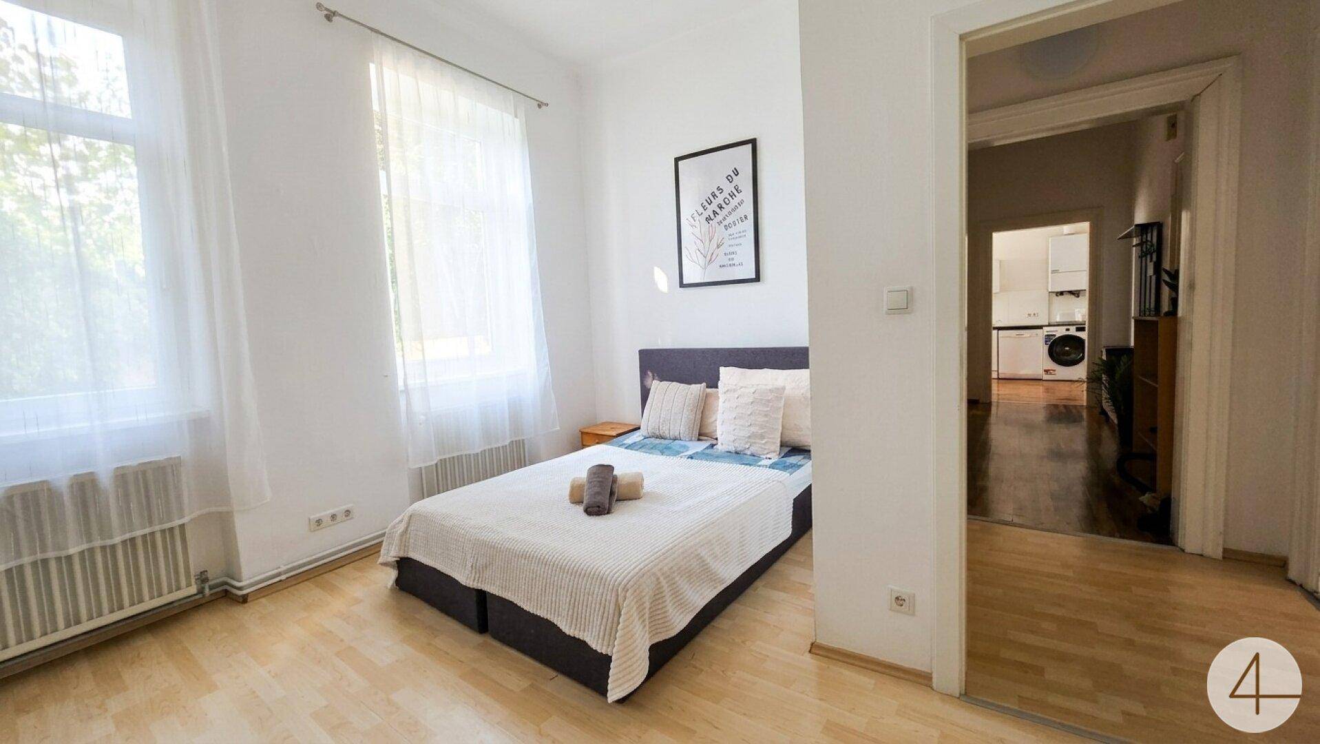 Helles Schlafzimmer mit Doppelbett, Laminatboden und großen Fenstern.