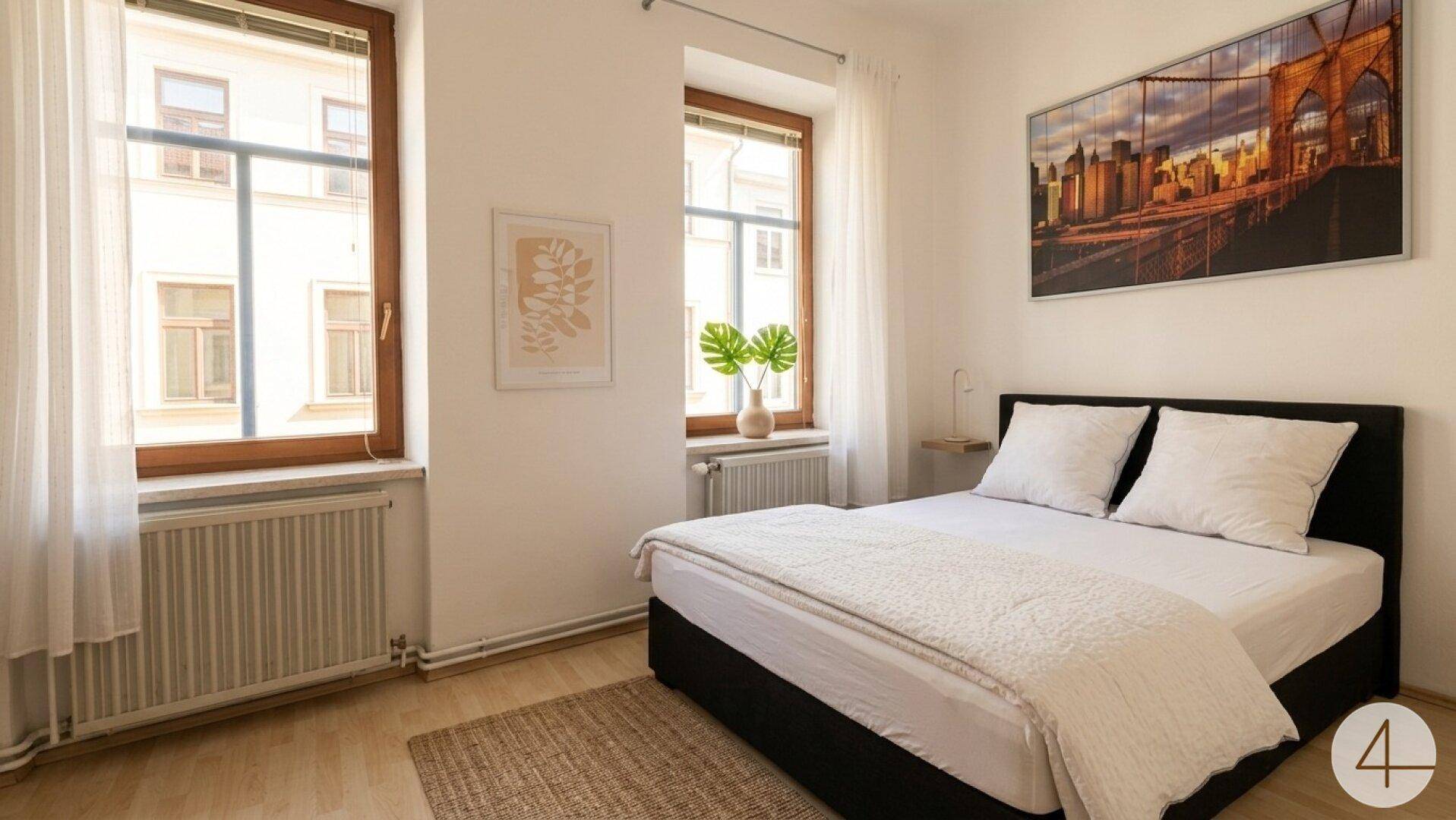 Helles Schlafzimmer mit Doppelbett, Holzboden und zwei großen Fenstern.