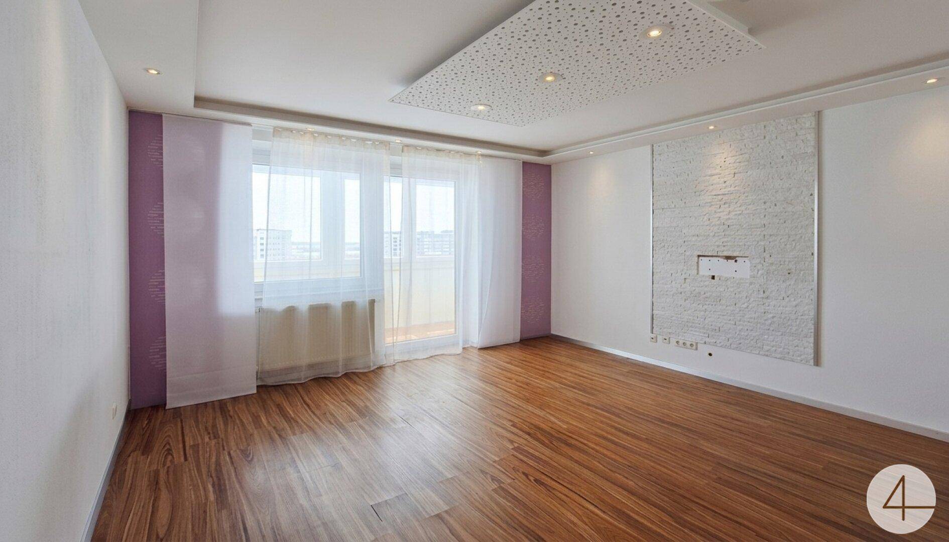 Heller Wohnraum mit Holzboden, großem Fenster und Zugang zu einem Balkon.