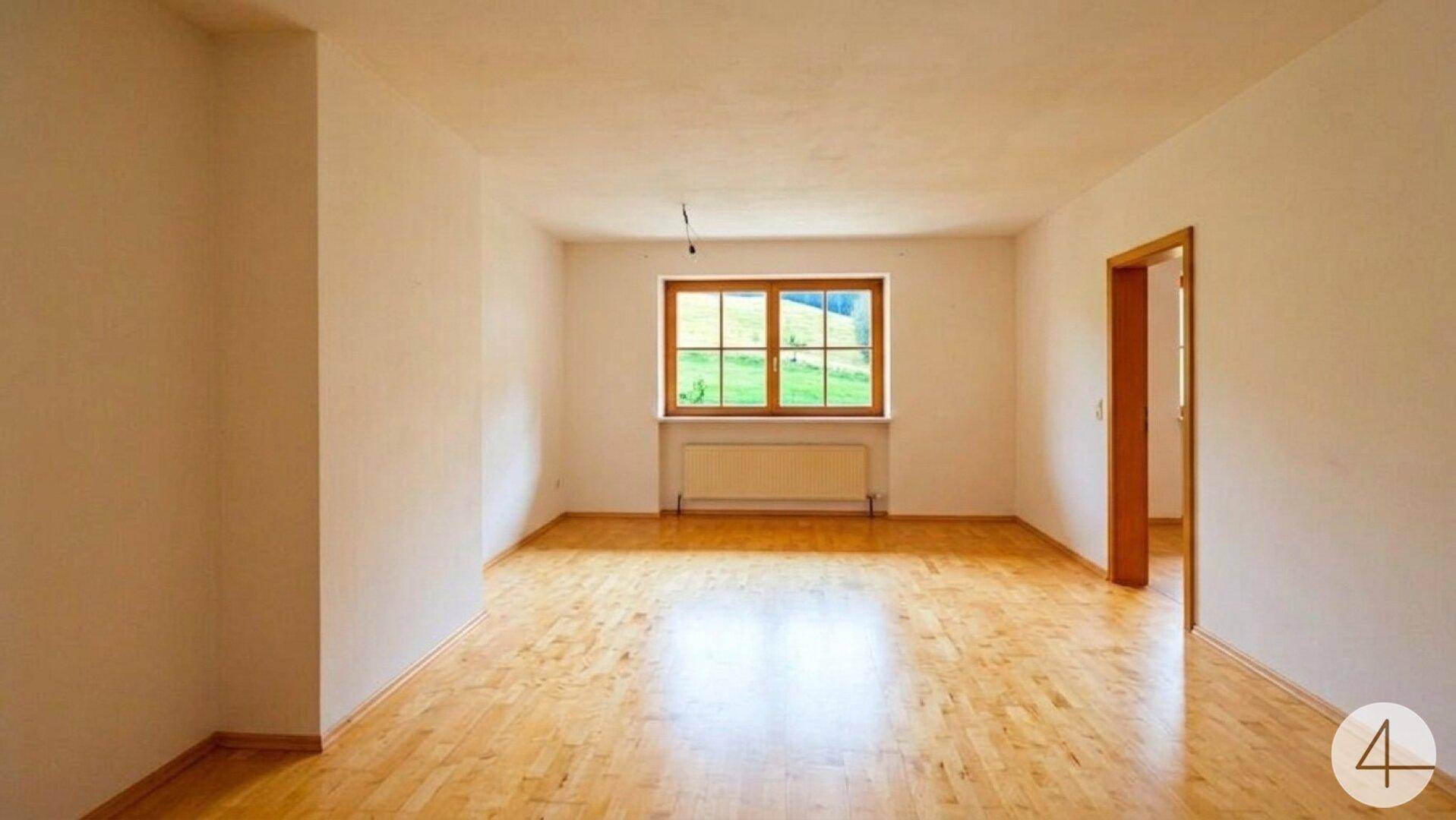 Heller, leerer Raum mit Holzboden und einem großen Fenster mit Blick ins Grüne.