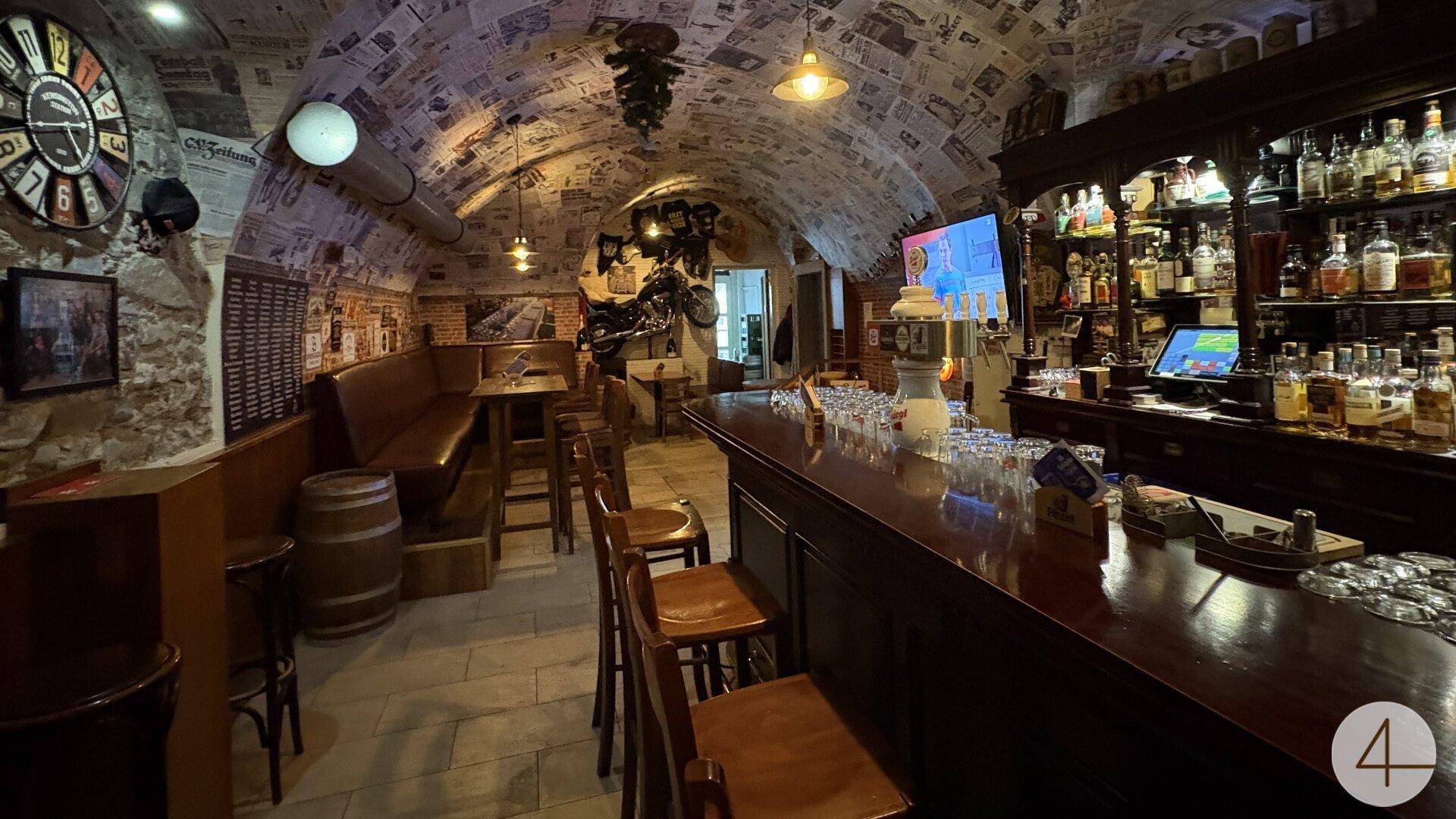 Gemütliche Bar mit langer Holztheke, reichhaltiger Getränkeauswahl und einzigartiger Zeitungs-Deckenverkleidung.