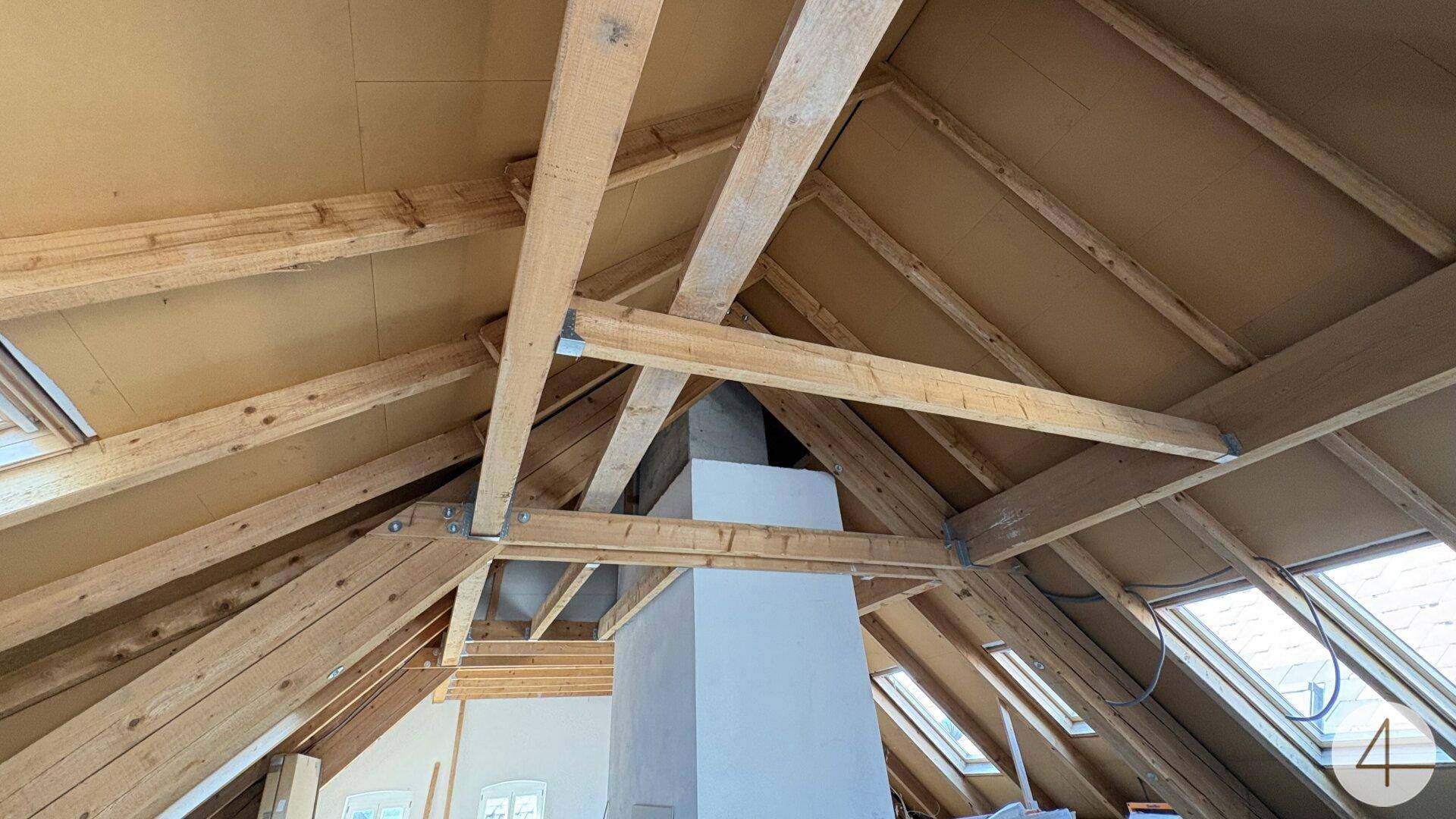 Dachboden im Rohbau mit sichtbarer Holzkonstruktion, Schornstein und mehreren Dachfenstern.
