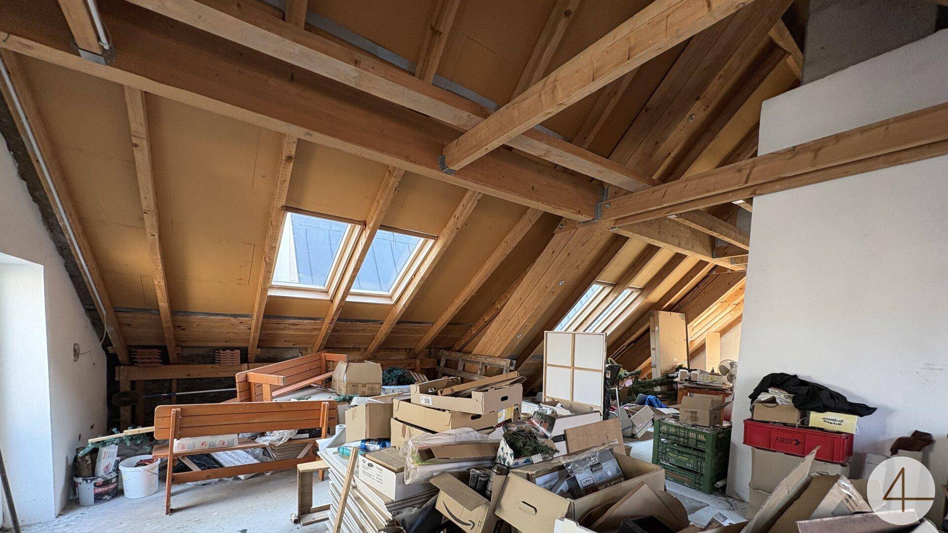 Unaufgeräumter Dachboden mit sichtbarer Holzkonstruktion, Dachfenstern und gelagerten Gegenständen.