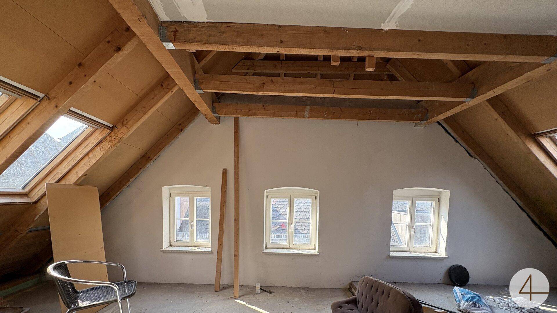 Teilweise ausgebauter Dachraum mit sichtbaren Holzbalken, Dachfenstern und drei kleinen Wandfenstern.