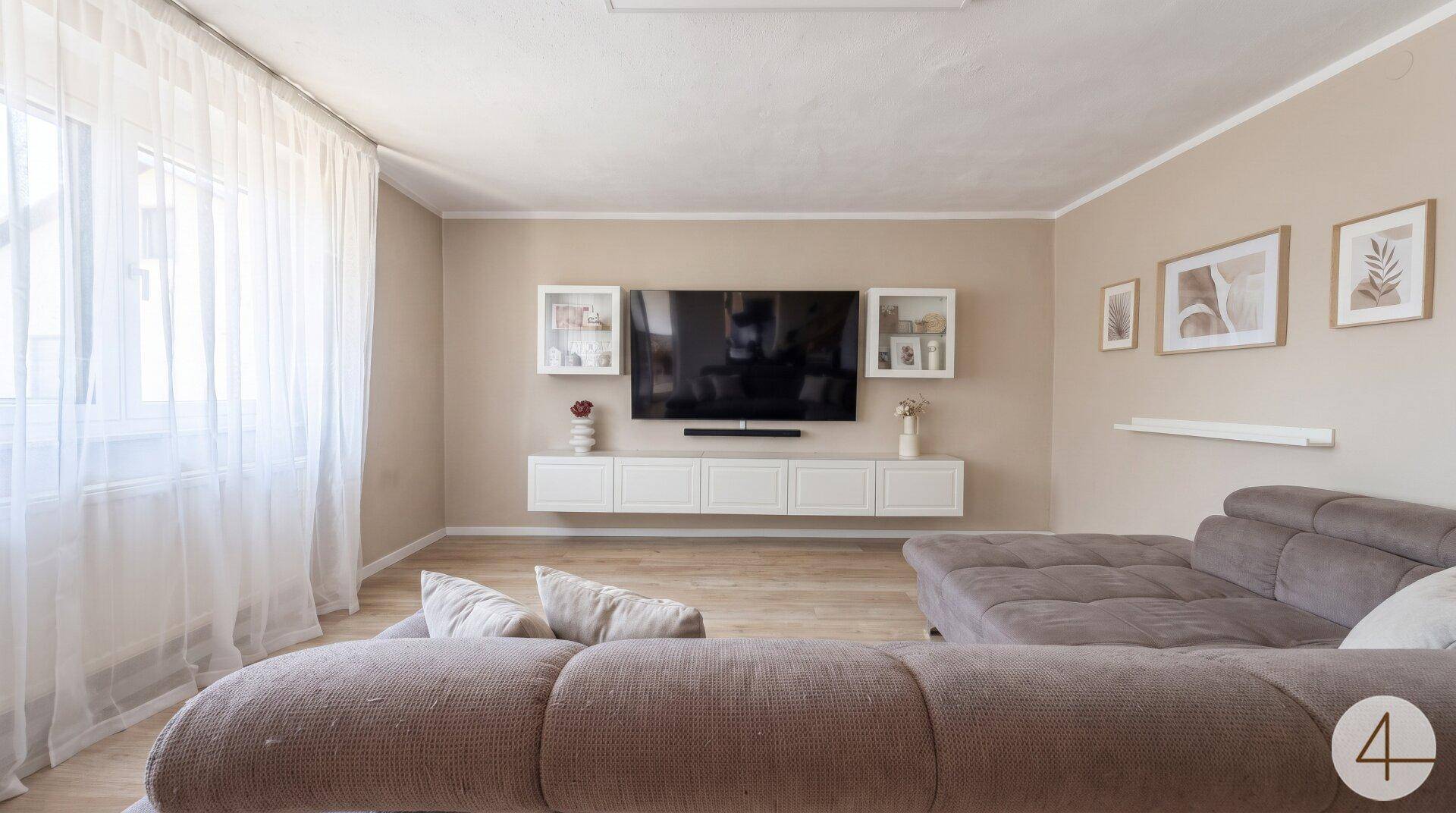 Helles Wohnzimmer mit beigem Sofa, großem Fernseher und Fenster mit Vorhängen.