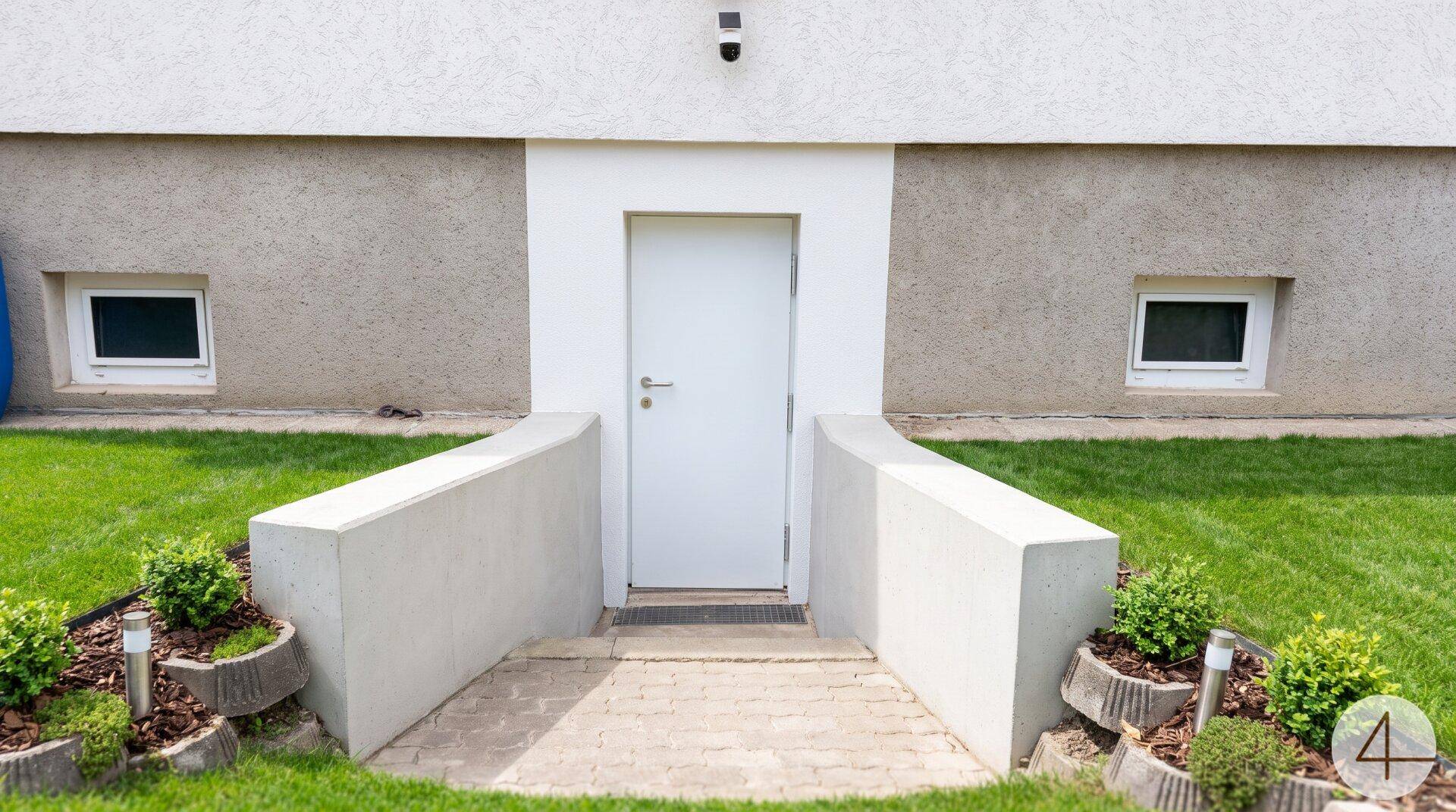 Eingang zum Kellergeschoss mit weißer Tür und kleinen Fenstern, umgeben von Rasen.