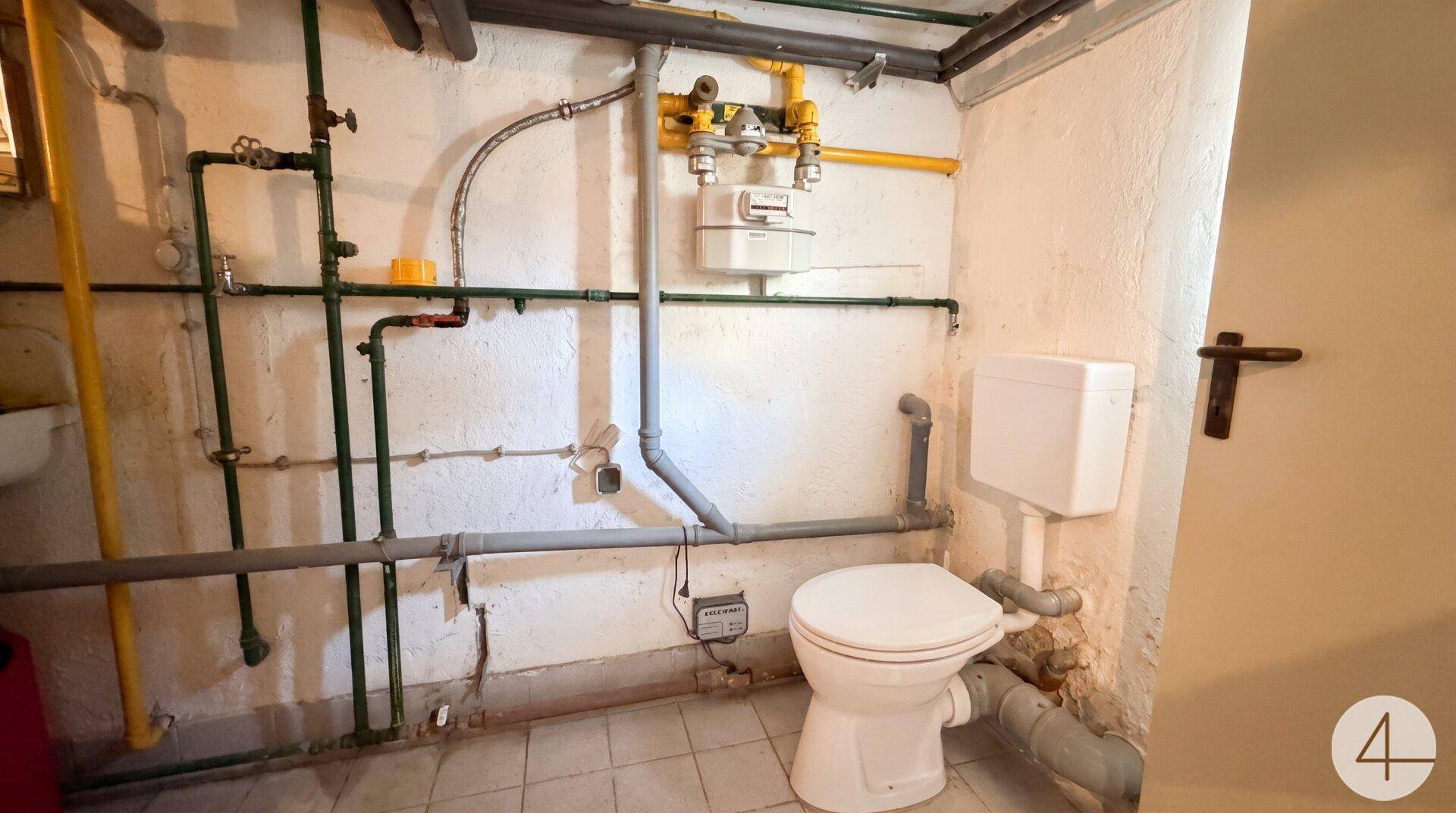 Einfache Toilette im Kellerbereich mit sichtbaren Installationen und Gaszähler.