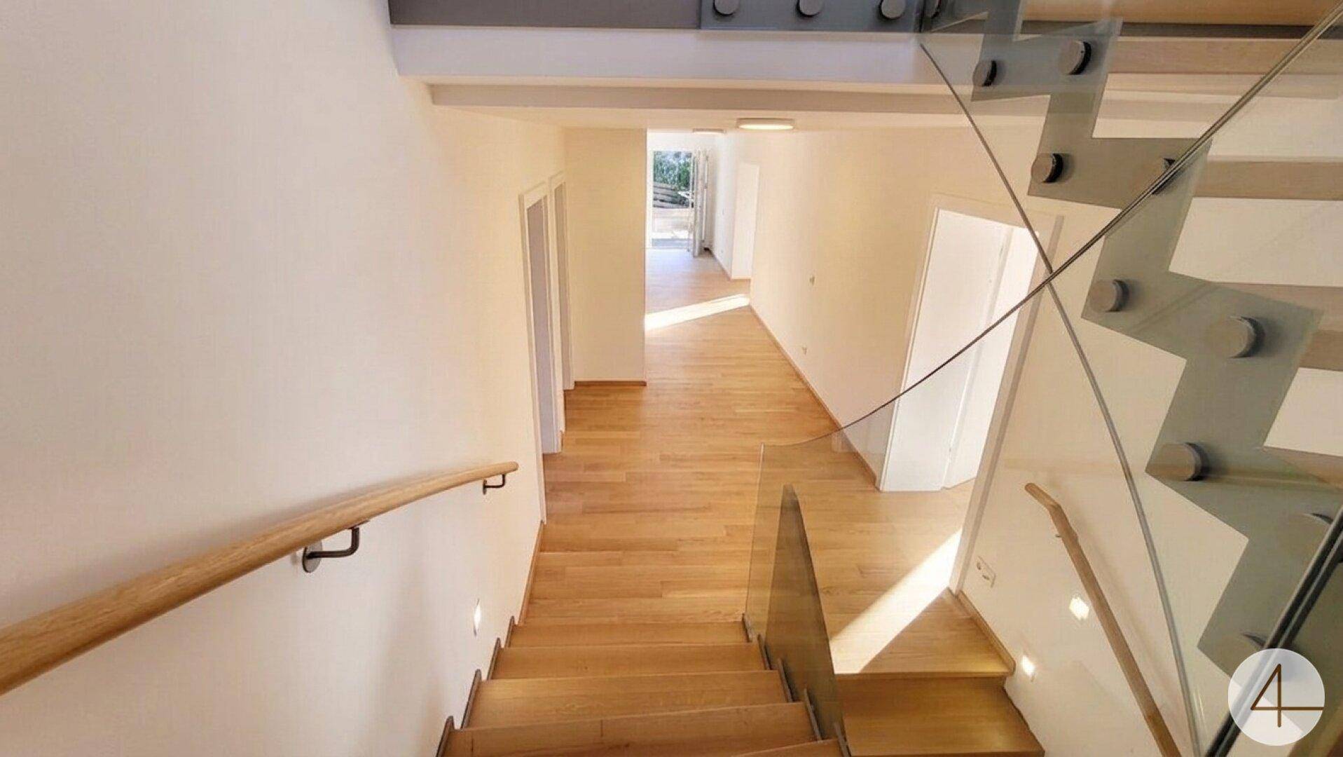 Moderner Treppenaufgang mit Glasgeländer und Holzstufen, der zu hellen Fluren führt.