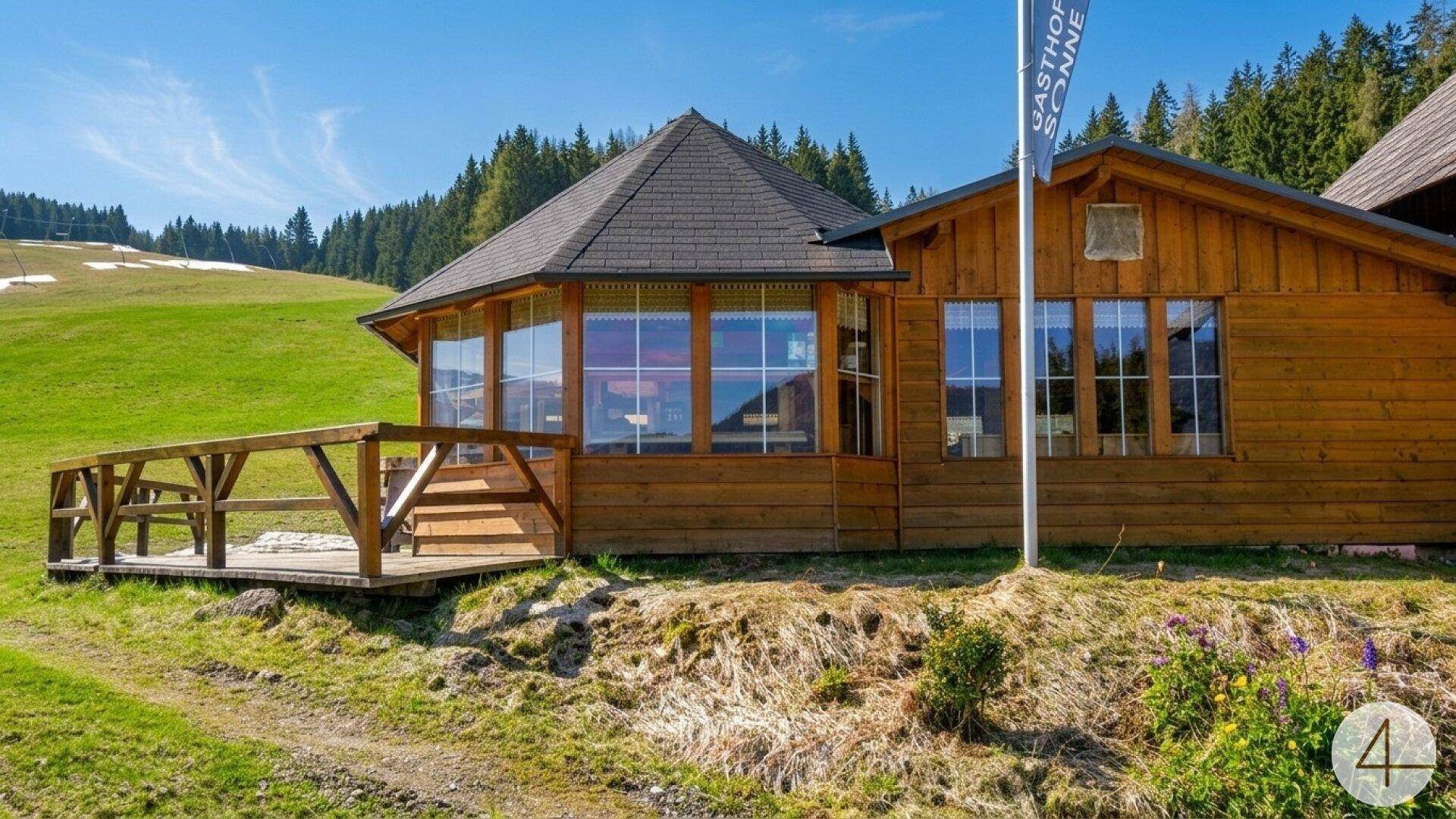 Holzgebäude mit großen Fenstern und einer Holzterrasse, umgeben von grünen Wiesen und einem schneebedeckten Hang.