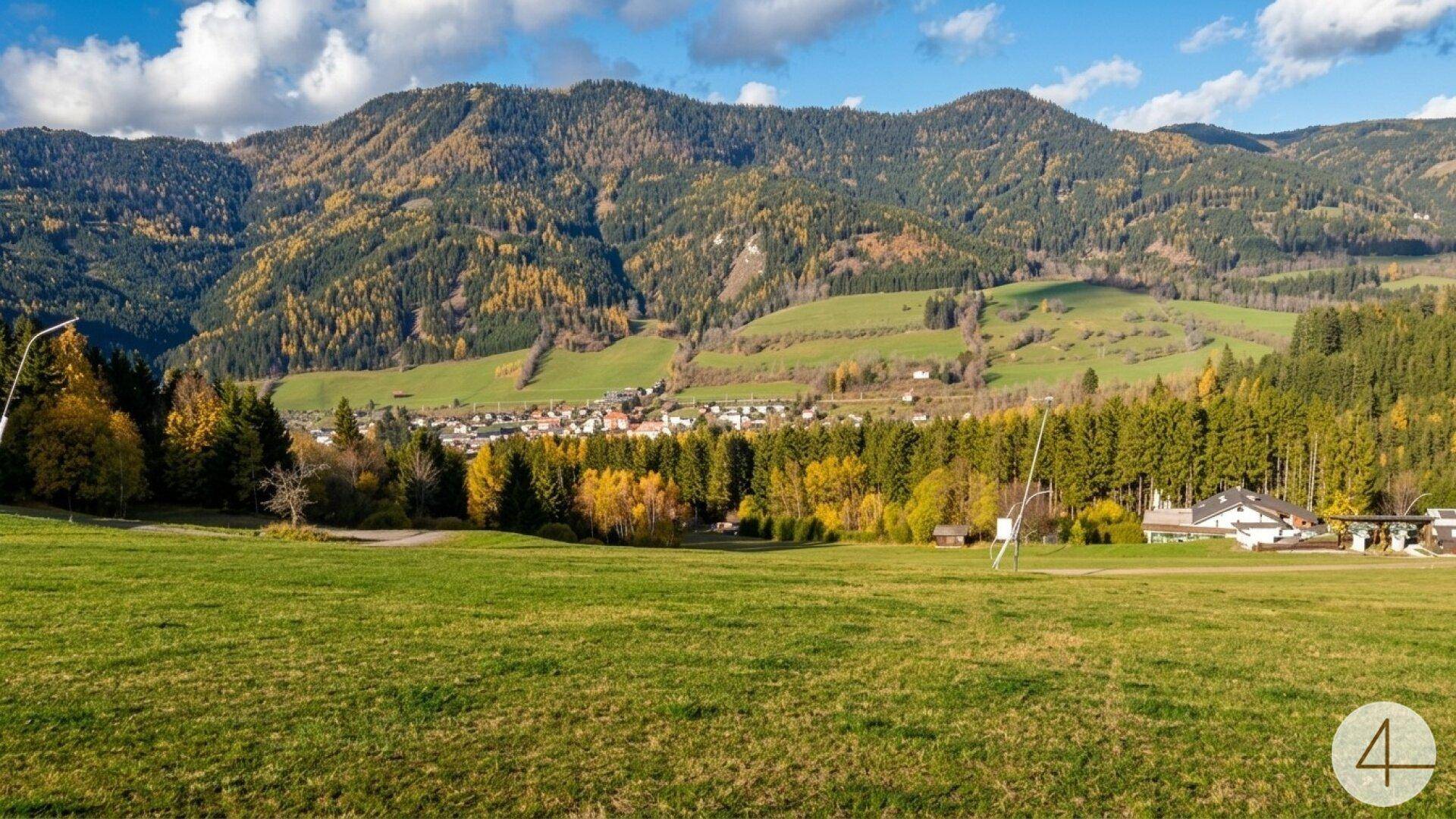 Panoramablick über eine grüne Hügellandschaft mit einem Dorf, umgeben von Wäldern und Bergen.