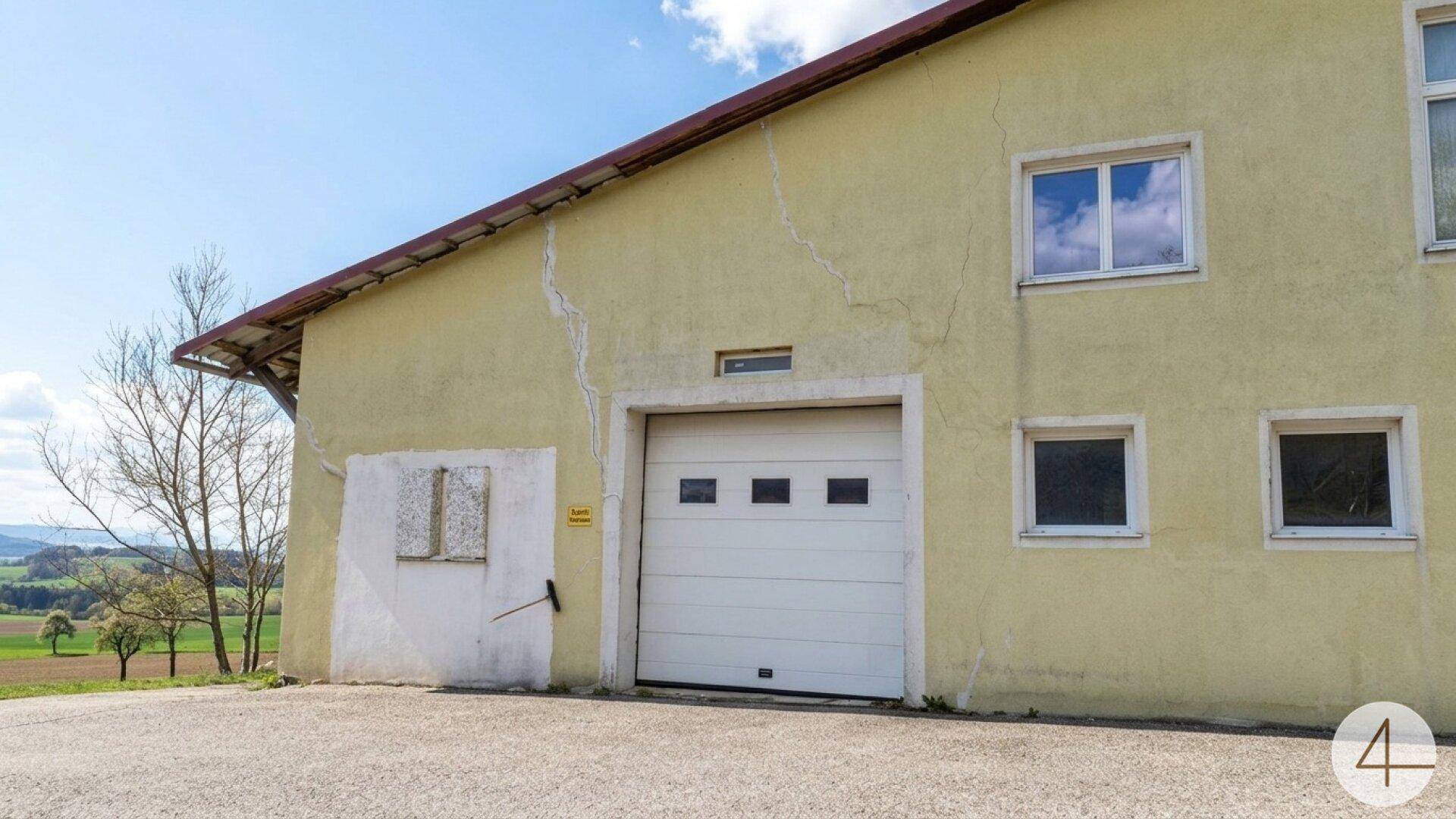 Fassade eines Gebäudes mit Garagentor und Rissen im Putz, umgeben von ländlicher Landschaft.
