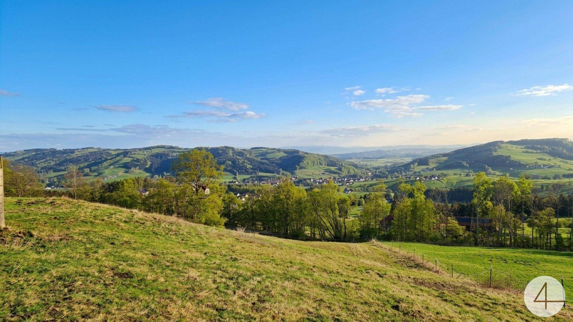 Panoramablick über sanfte grüne Hügel, Wälder und ein entferntes Dorf unter blauem Himmel.
