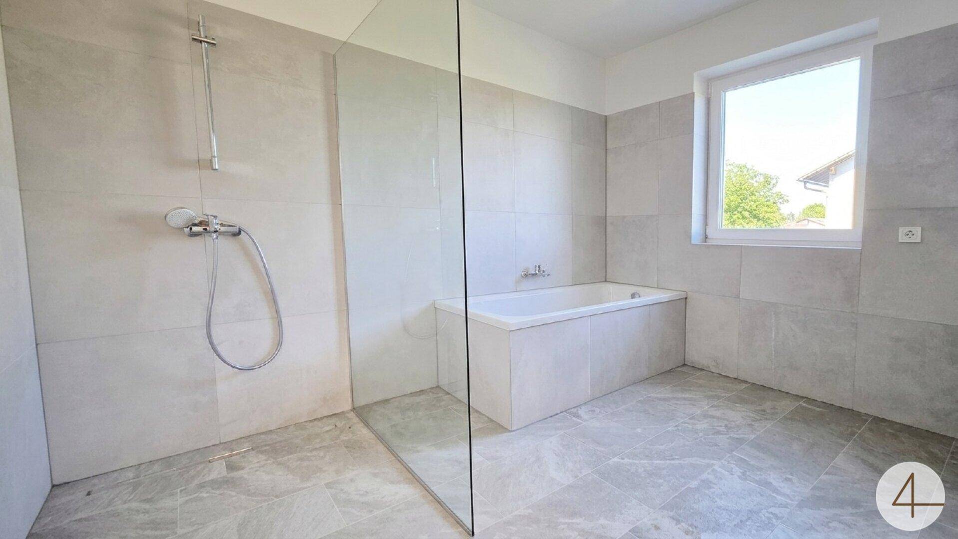Modernes Badezimmer mit grauen Fliesen, einer begehbaren Dusche und einer Badewanne unter dem Fenster.