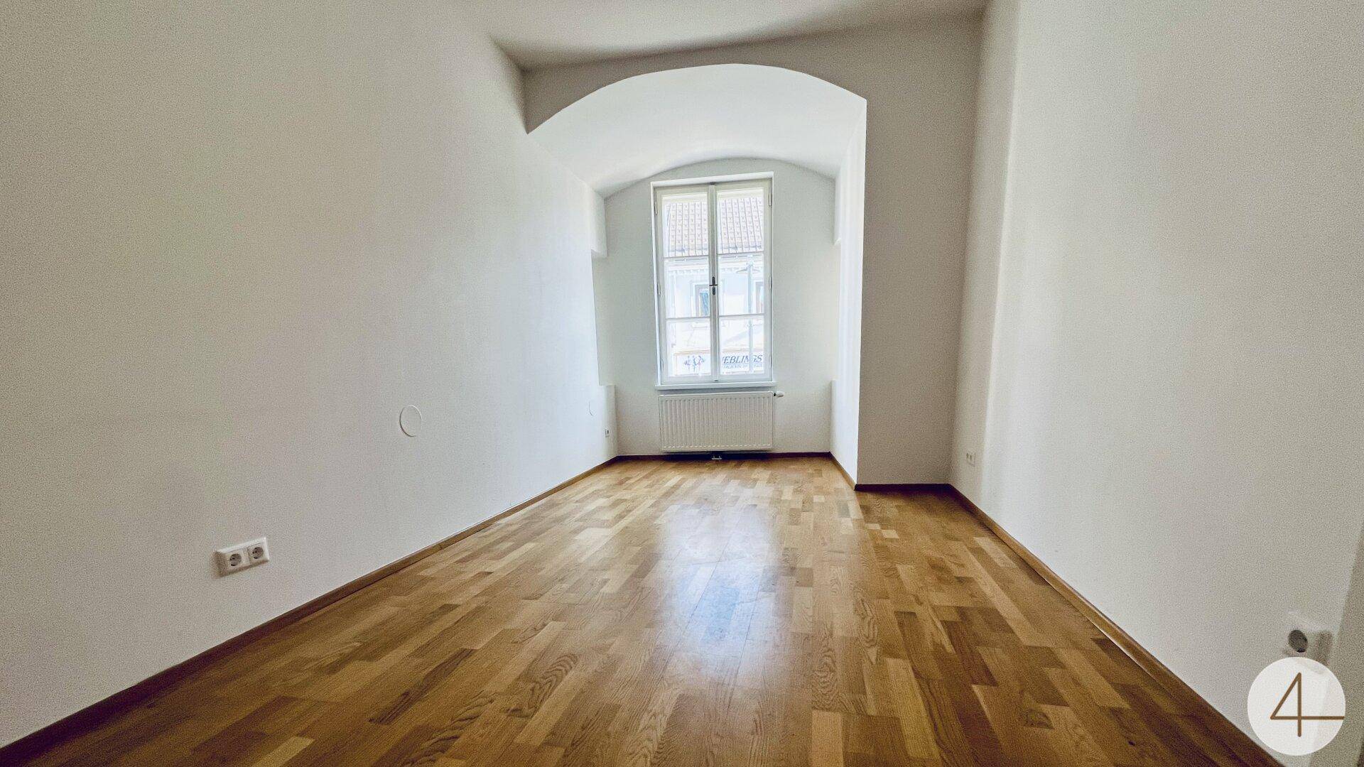 Ruhiges Zimmer mit Holzboden, einem Fenster und einem Heizkörper unter einem Bogen.