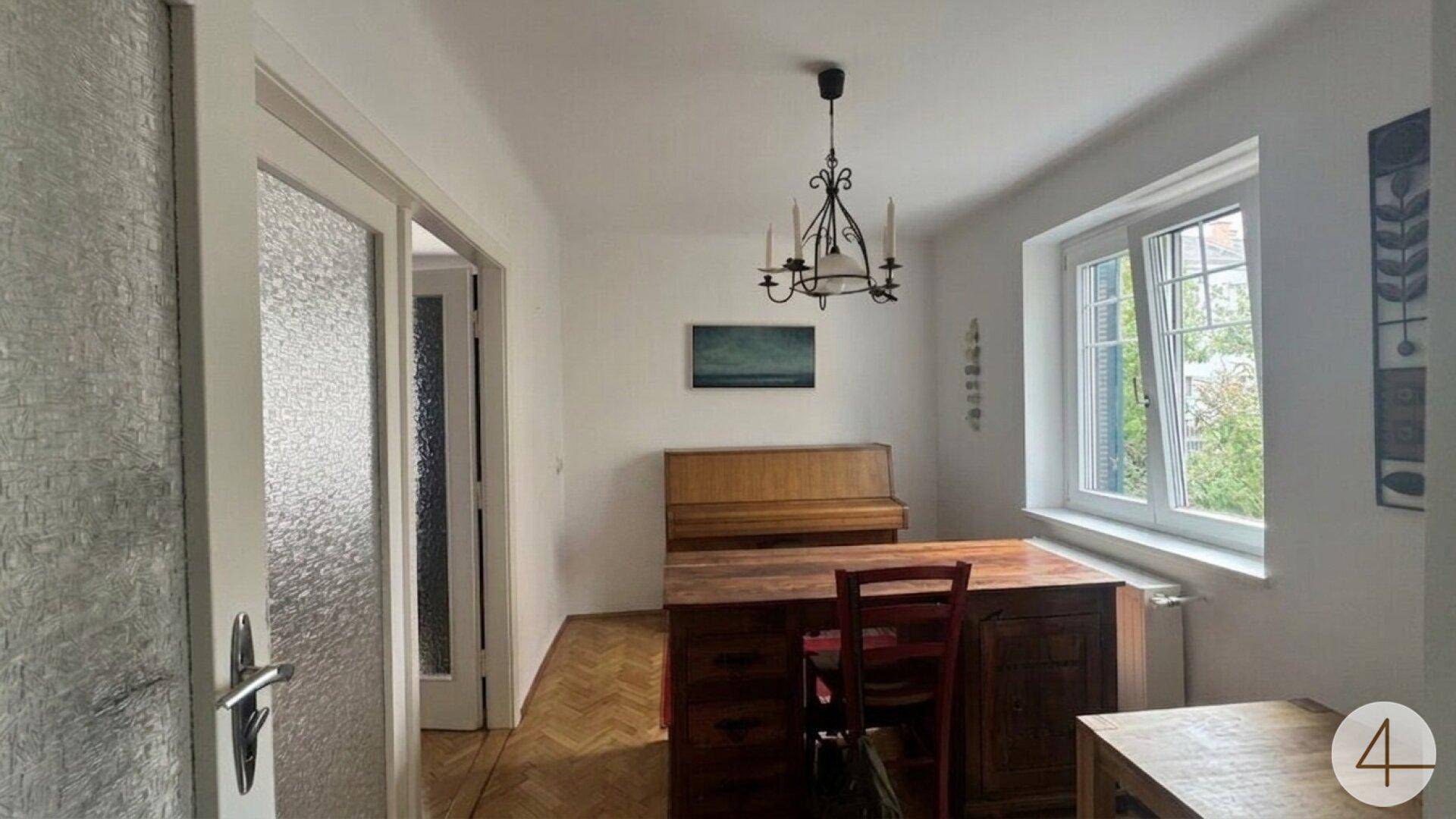 Helles Zimmer mit Holzboden, Schreibtisch und einem Klavier am Fenster.