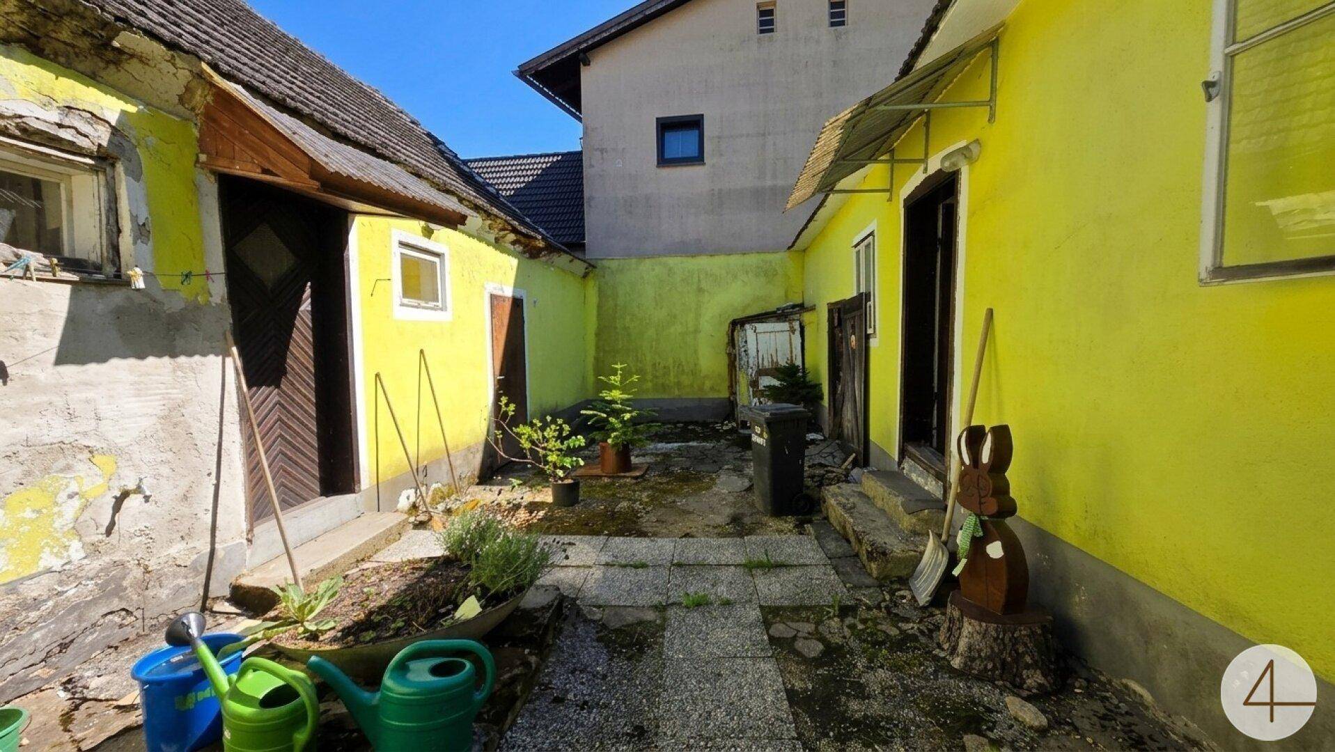 Hinterhof mit gelb gestrichenen Wänden, unebenem Boden und verschiedenen Gartengeräten.