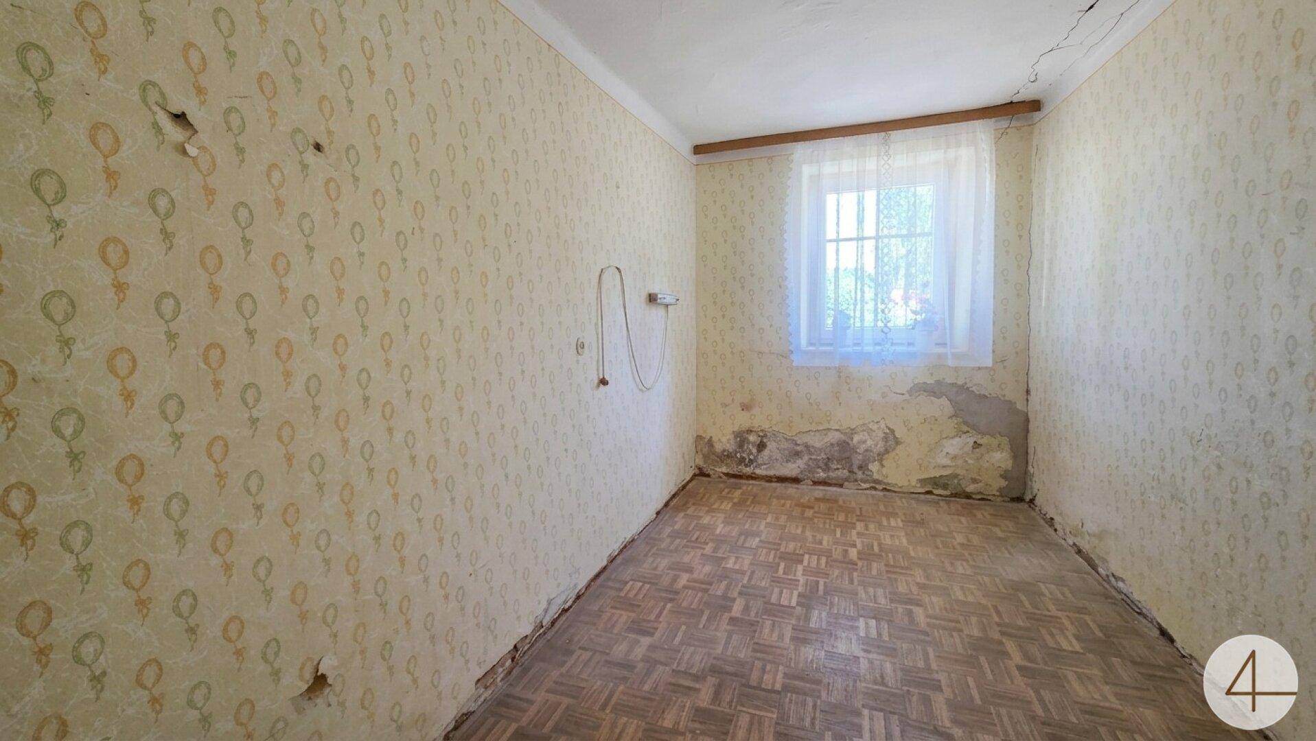 Kleines Zimmer mit abgenutzter Tapete, einem Fenster und sichtbaren Feuchtigkeitsschäden.