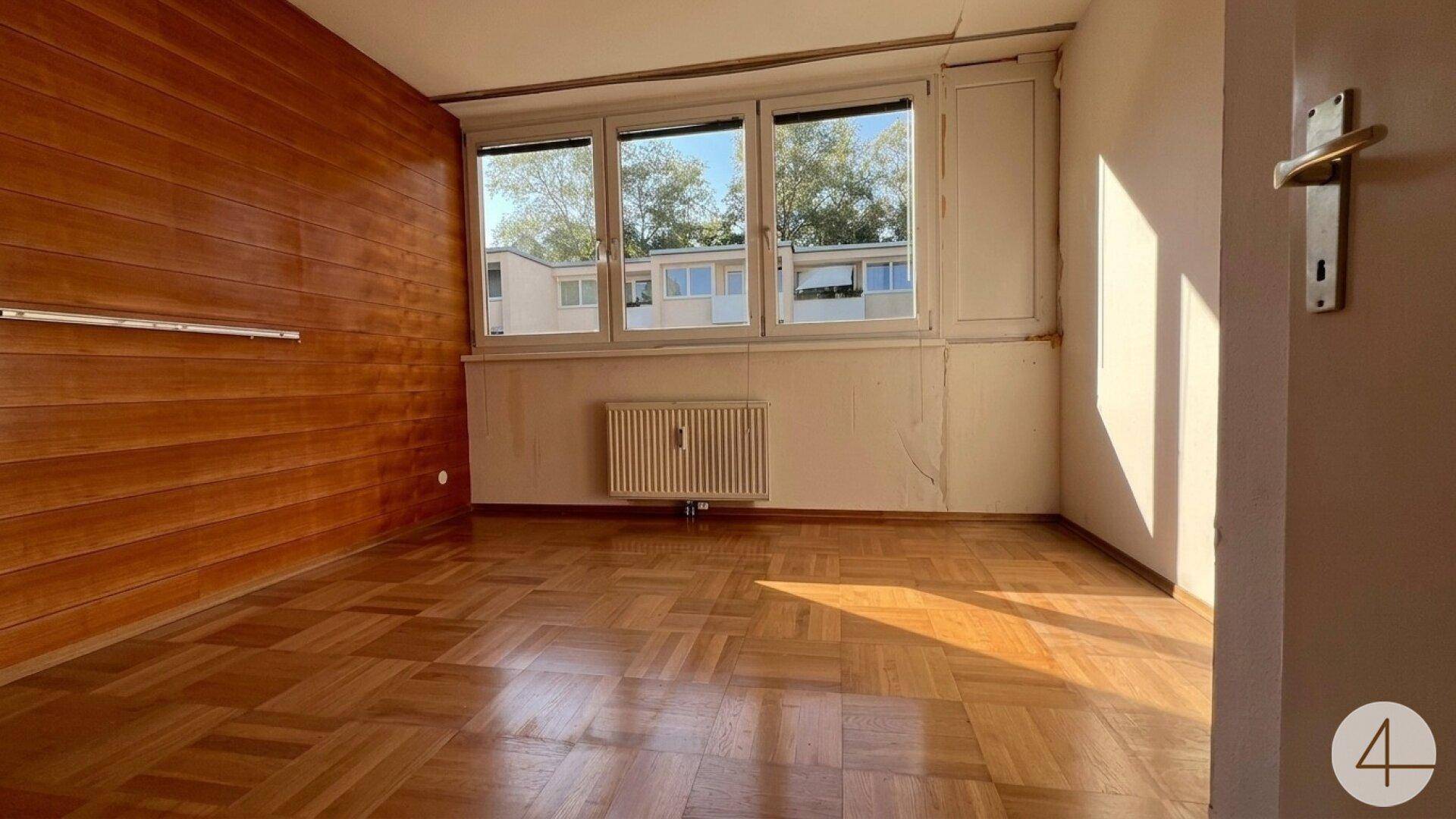 Geräumiges Zimmer mit Holzvertäfelung, Parkettboden und großen Fenstern mit Blick ins Grüne.