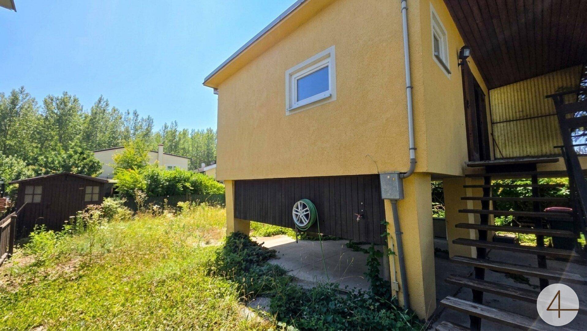 Seitenansicht des gelben Hauses mit Außentreppe und einem ungepflegten Gartenbereich.