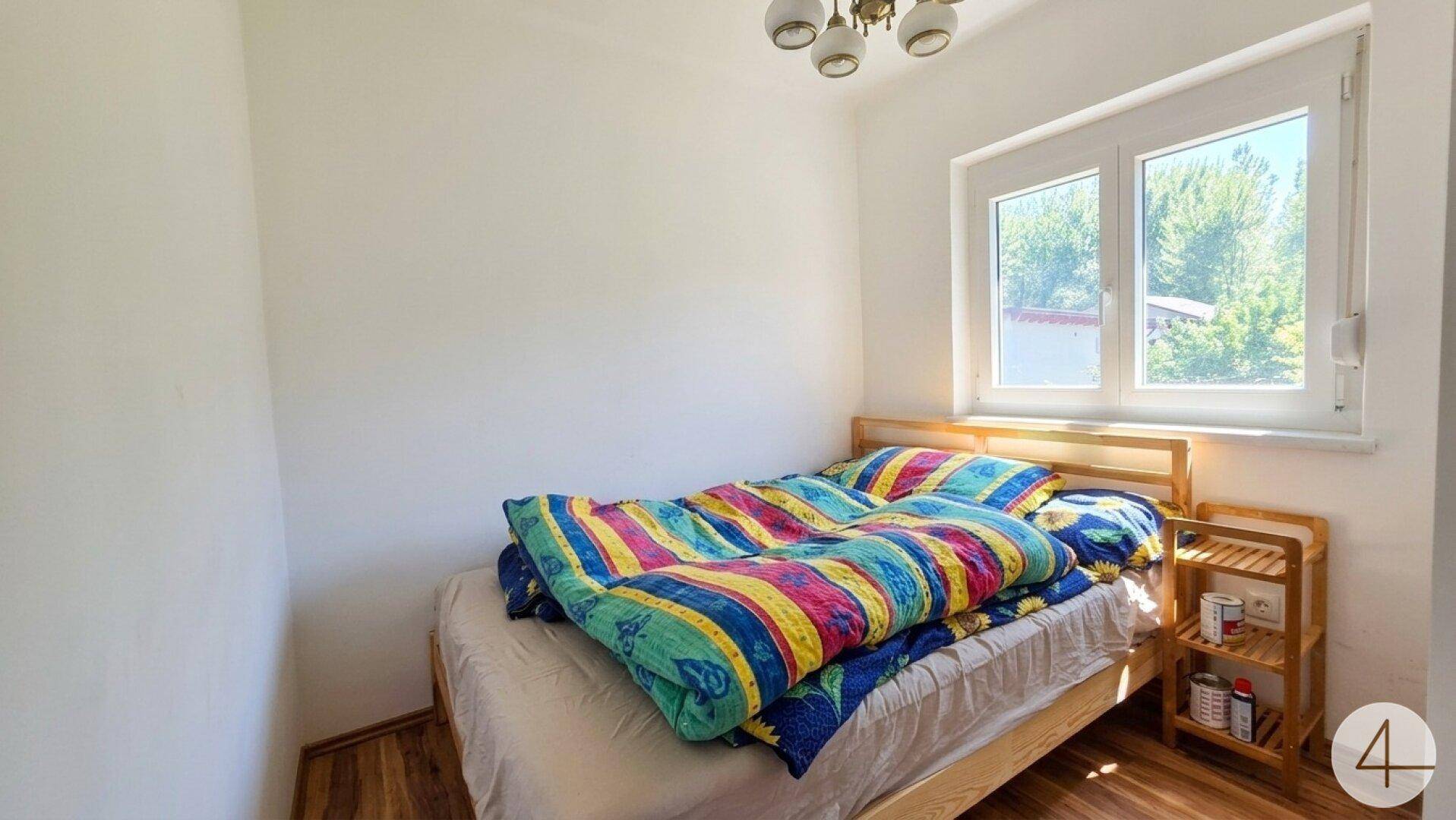 Helles Schlafzimmer mit Holzbett, bunten Decken und einem Fenster mit Blick ins Grüne.