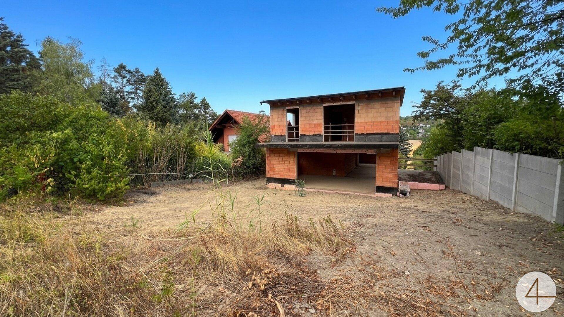 Rohbau eines Hauses mit unverputzten Ziegelwänden und umgebender Vegetation auf einem Grundstück.