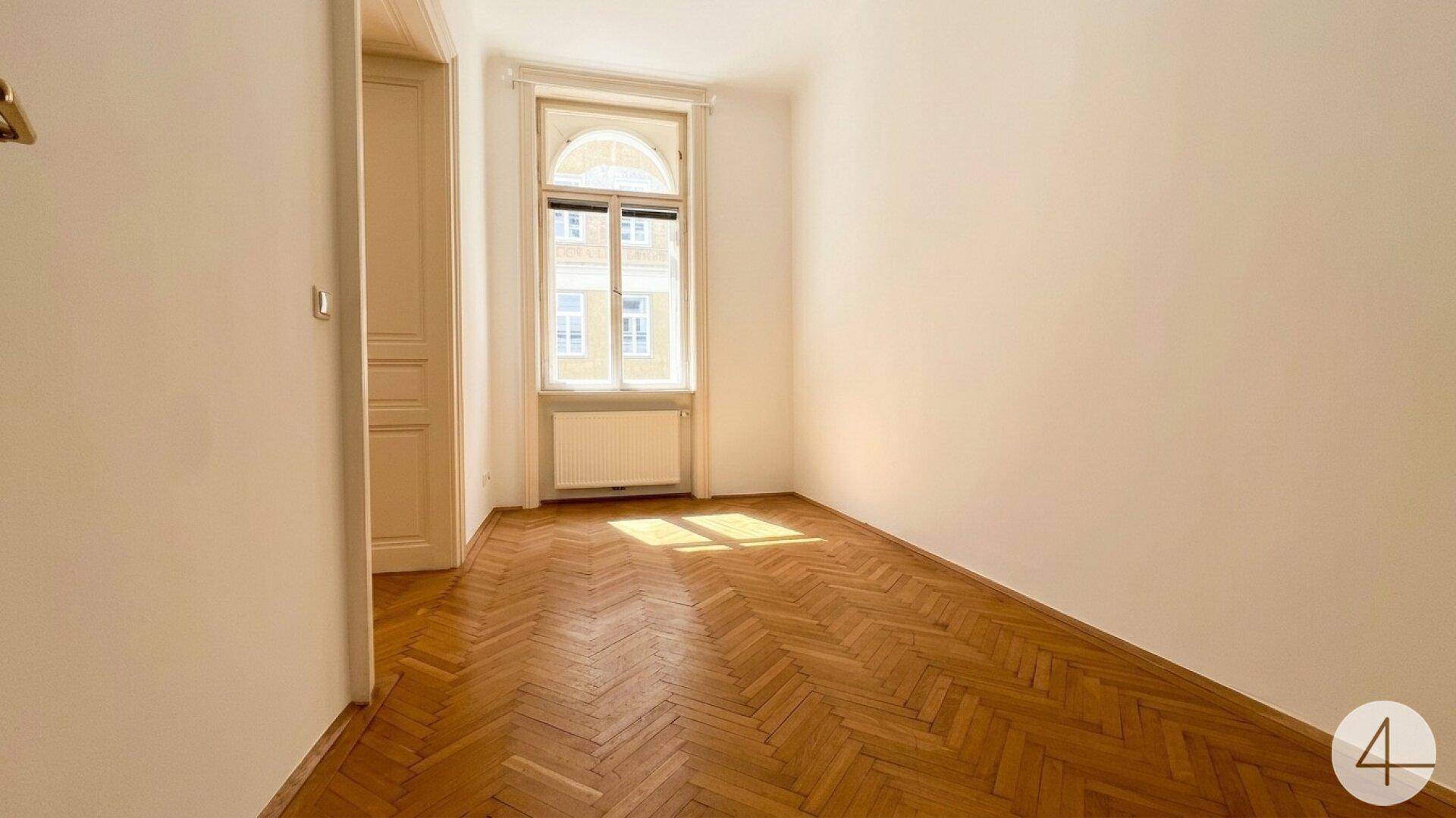 Gemütliches Zimmer mit Fischgrätparkett und einem großen Fenster, das den Raum hell erscheinen lässt.