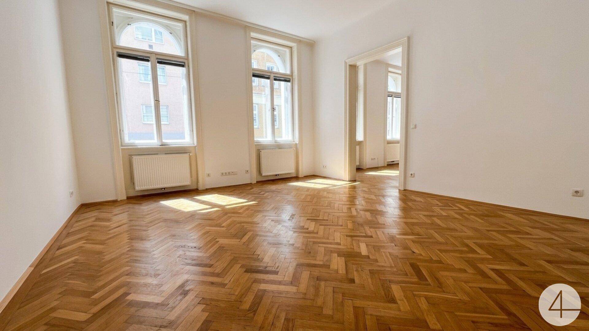 Heller Wohnraum mit elegantem Fischgrätparkett und hohen Fenstern, der einen offenen Übergang zu weiteren Zimmern bietet.