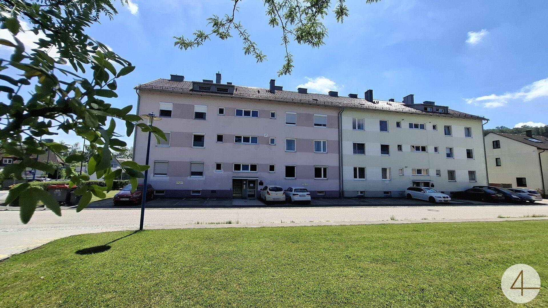 Mehrfamilienhaus mit heller Fassade, Satteldach und Grünfläche im Vordergrund unter blauem Himmel.