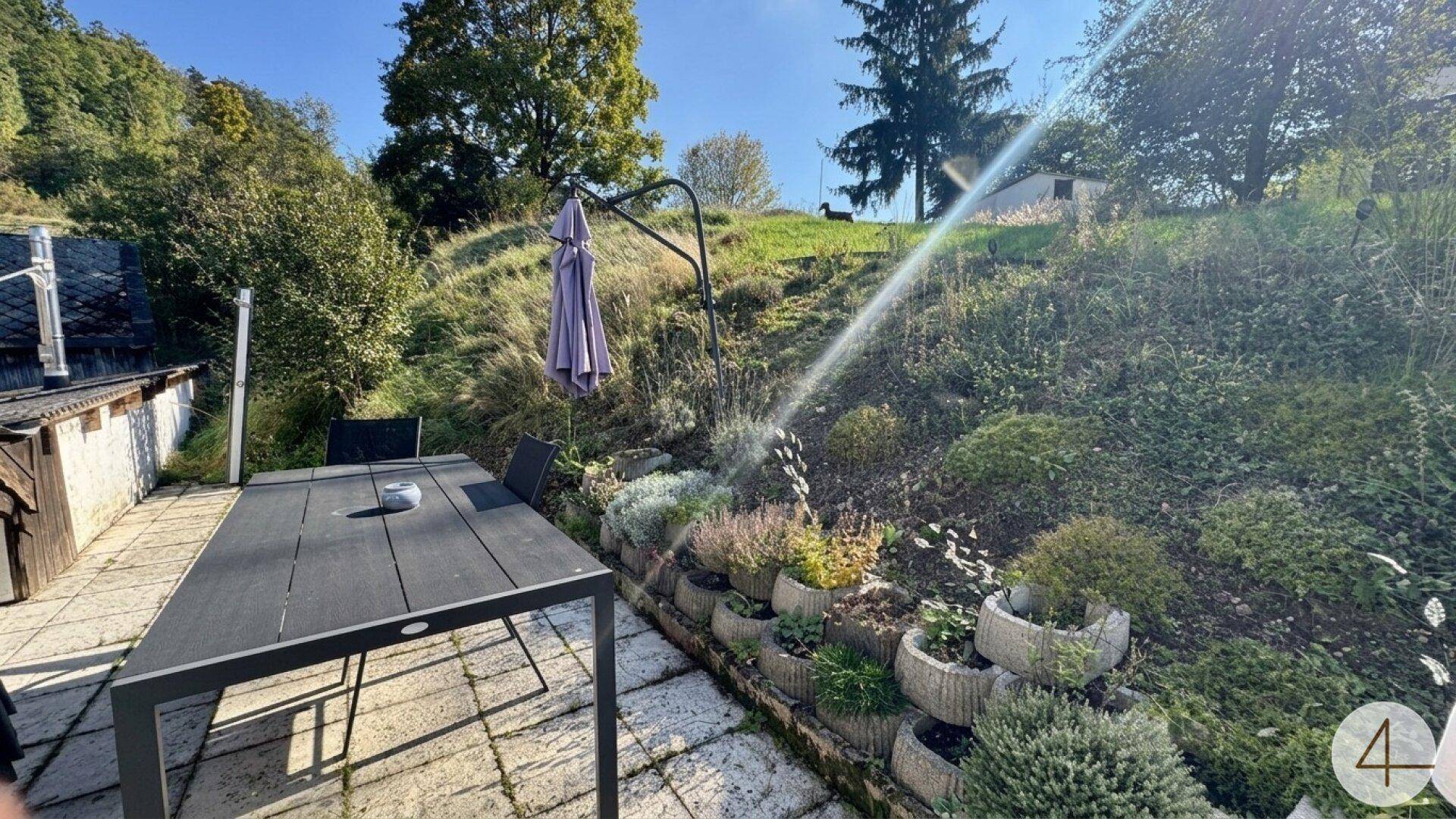 Gepflasterte Terrasse mit Esstisch und Stühlen, umgeben von bepflanzten Beeten.