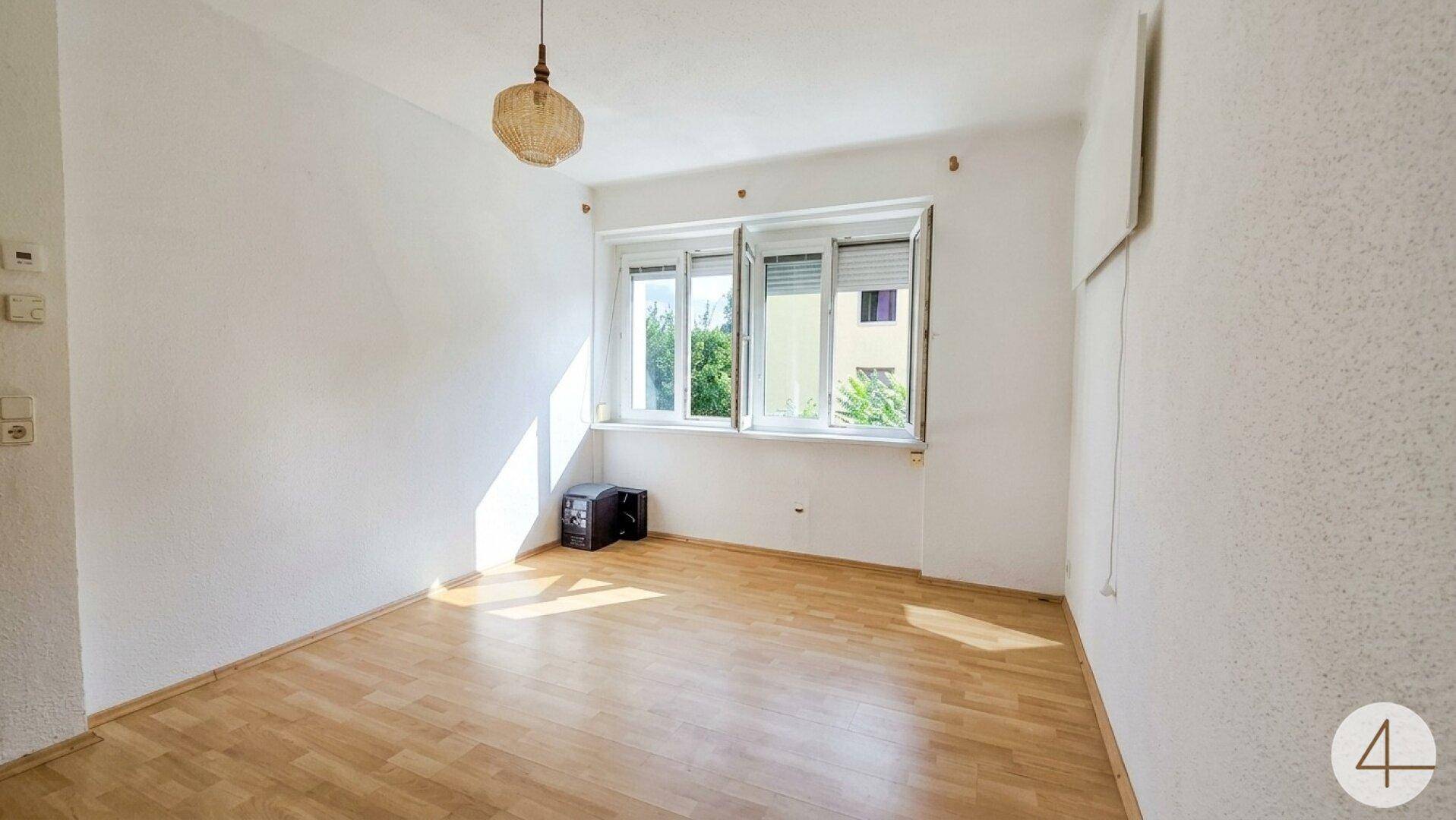 Helles, leeres Zimmer mit Laminatboden und Fenster mit Blick ins Grüne.