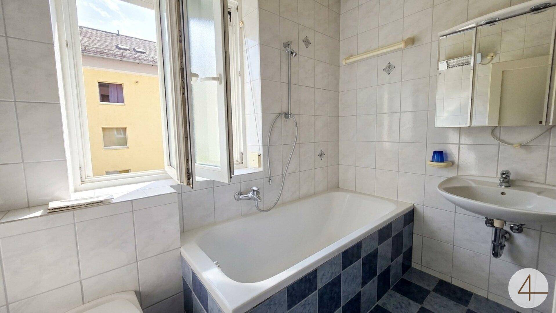 Badezimmer mit Badewanne, Dusche und Fenster, ausgestattet mit hellen Fliesen.