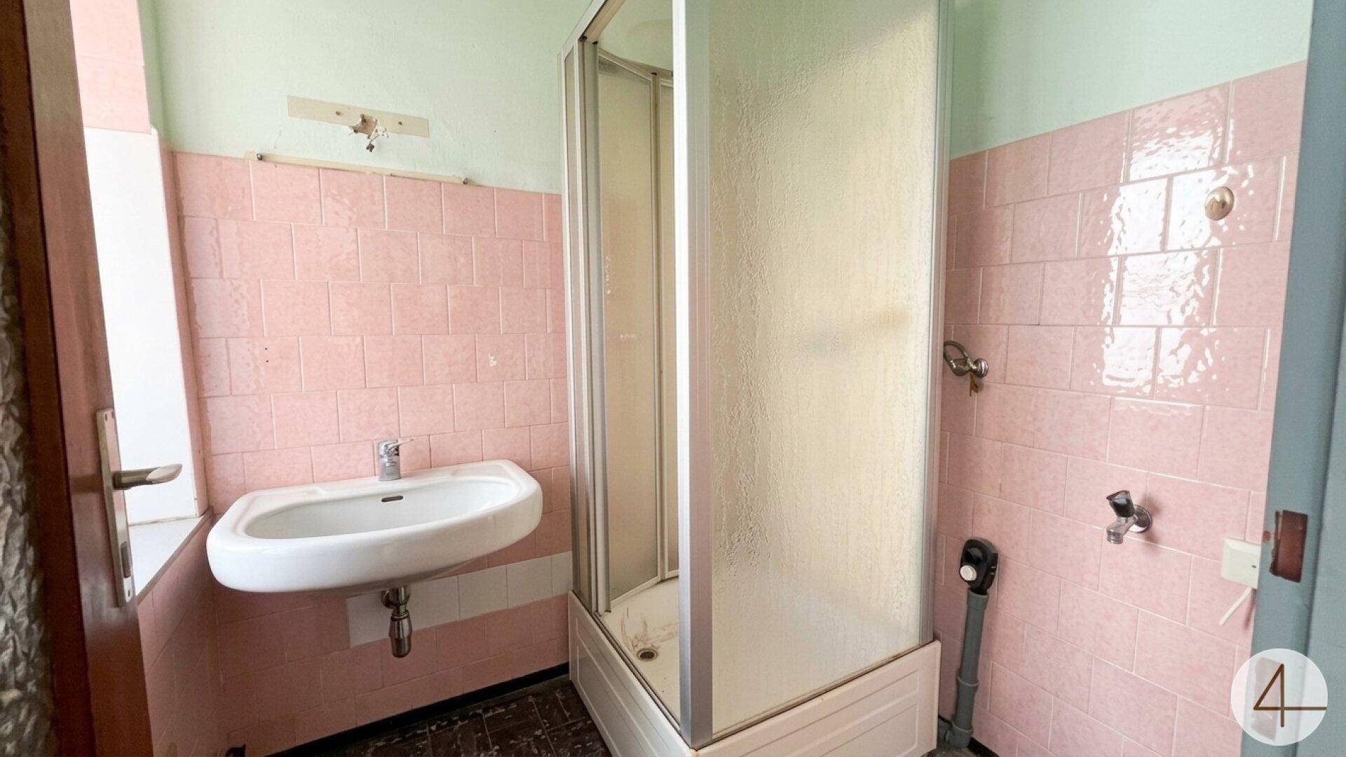 Veraltetes Badezimmer mit rosa Fliesen, Einzelwaschbecken und Duschkabine, renovierungsbedürftig.