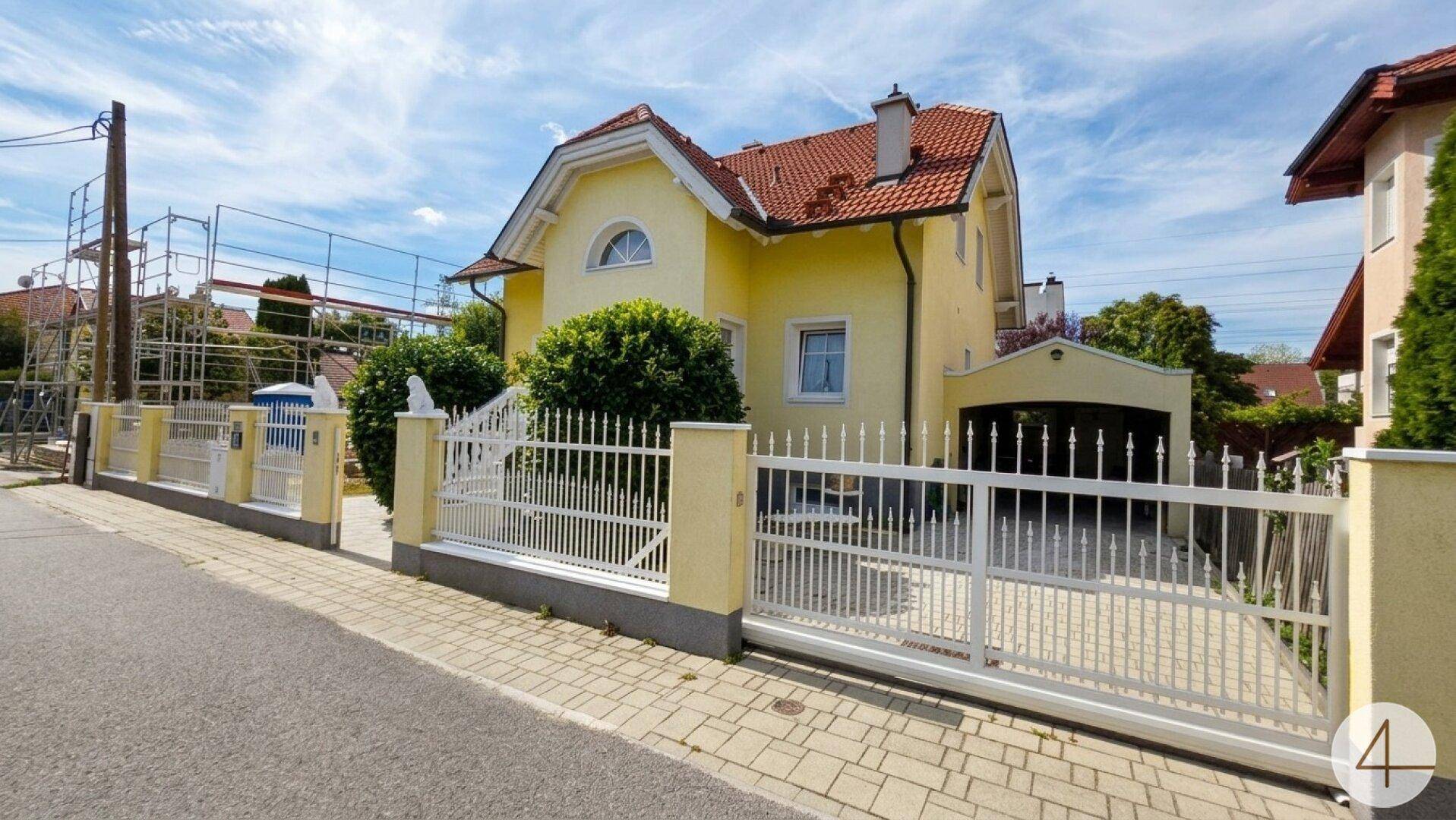 Seitenansicht des Hauses mit gelber Fassade, weißem Zaun, Einfahrtstor und Carport.