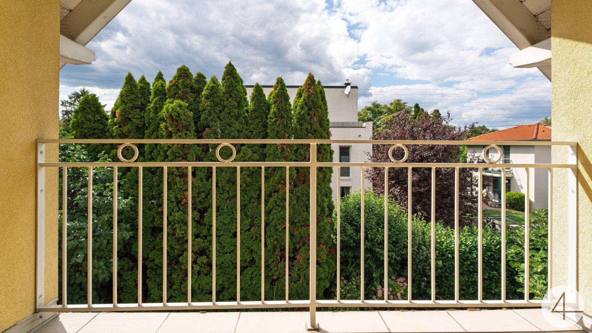 Balkon mit Ausblick auf grüne Bäume und entfernte Gebäude unter bewölktem Himmel.
