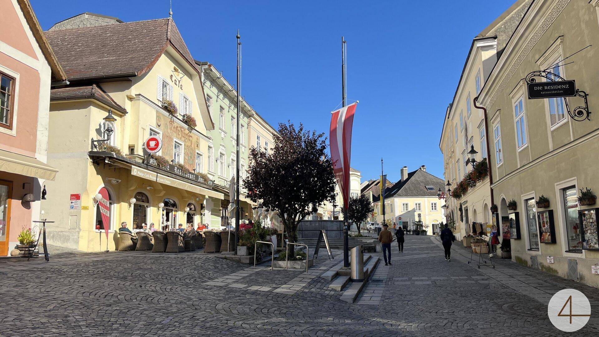 Belebter Marktplatz mit historischen Gebäuden, Außengastronomie und blauem Himmel an einem sonnigen Tag.