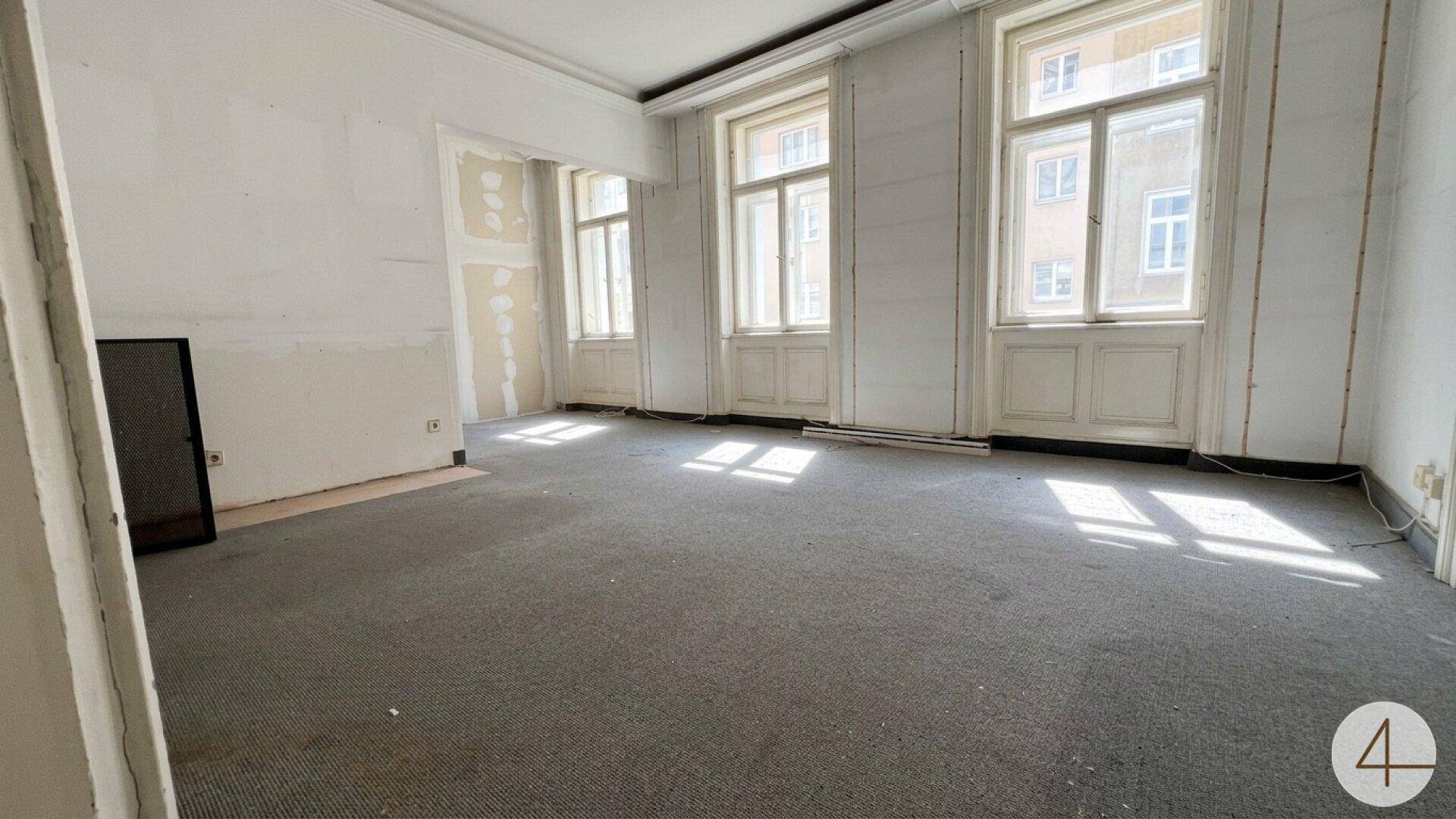 Geräumiger, heller Raum mit Teppichboden und hohen Fenstern, teilweise renovierungsbedürftig.