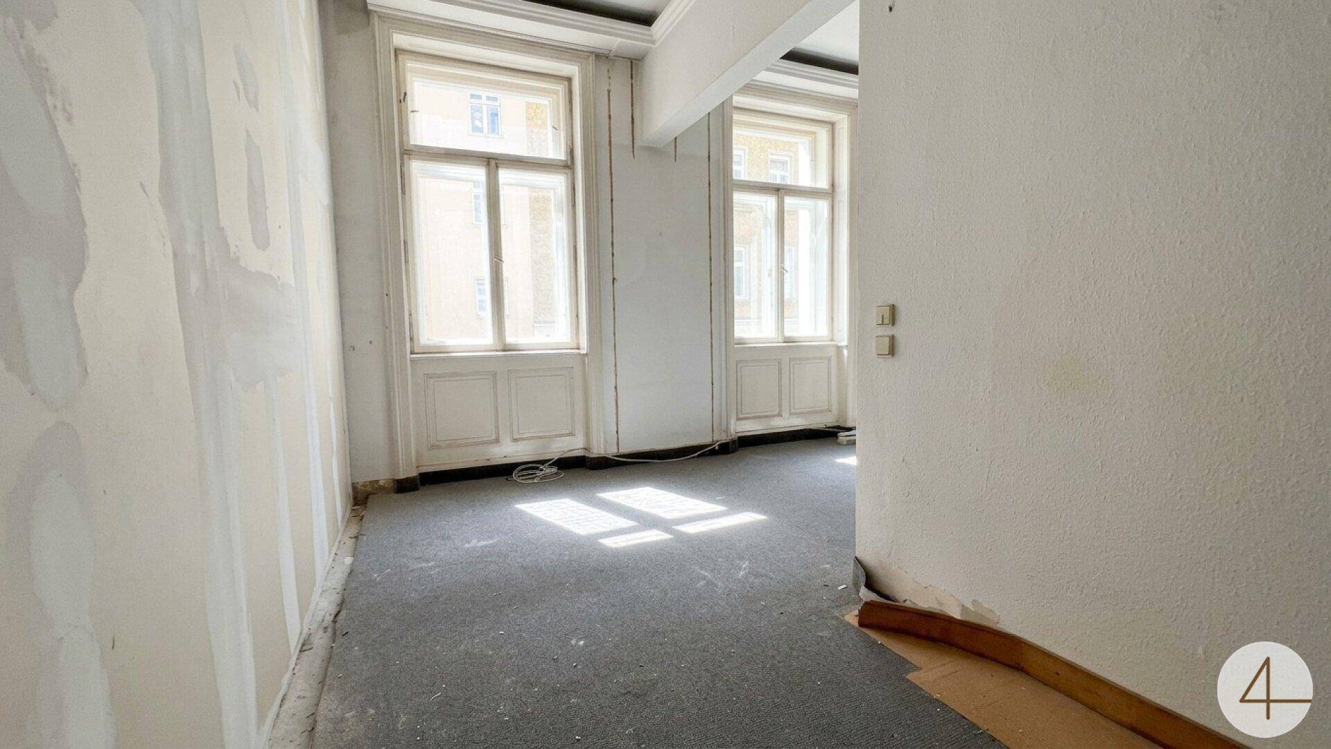 Heller Raum mit Teppichboden und hohen Fenstern, mit Spuren früherer Renovierungsarbeiten.