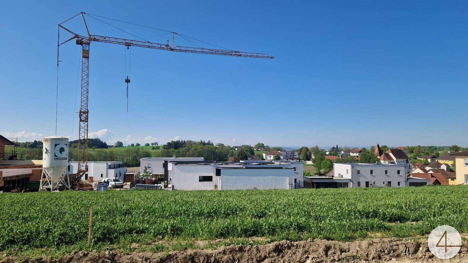 Baustelle mit Kran im Vordergrund, umgeben von einem Feld und Wohnhäusern in der Ferne unter blauem Himmel.