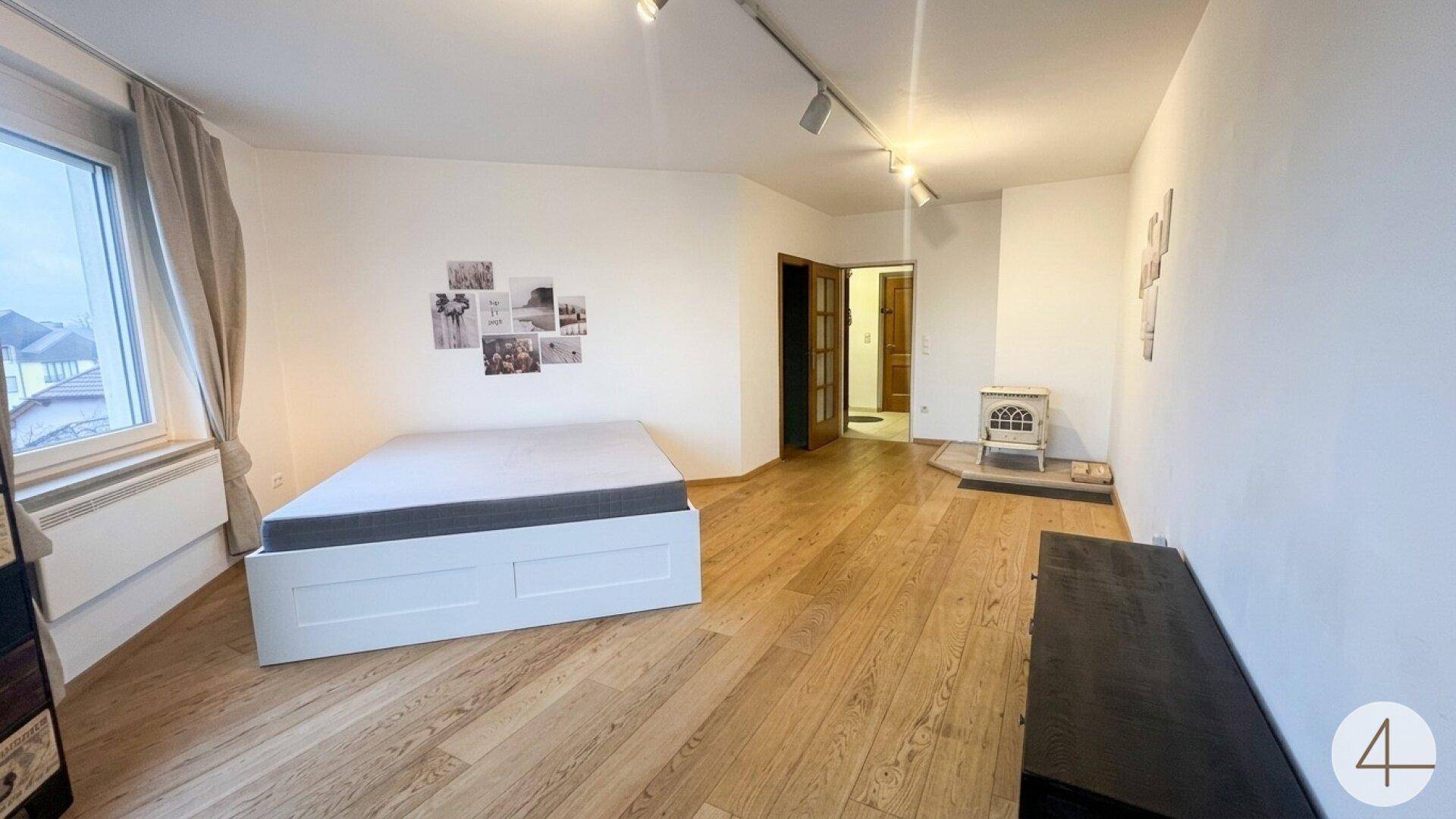 Geräumiges Wohnzimmer mit Holzboden, einem Bett und einem traditionellen Ofen, hell und einladend.