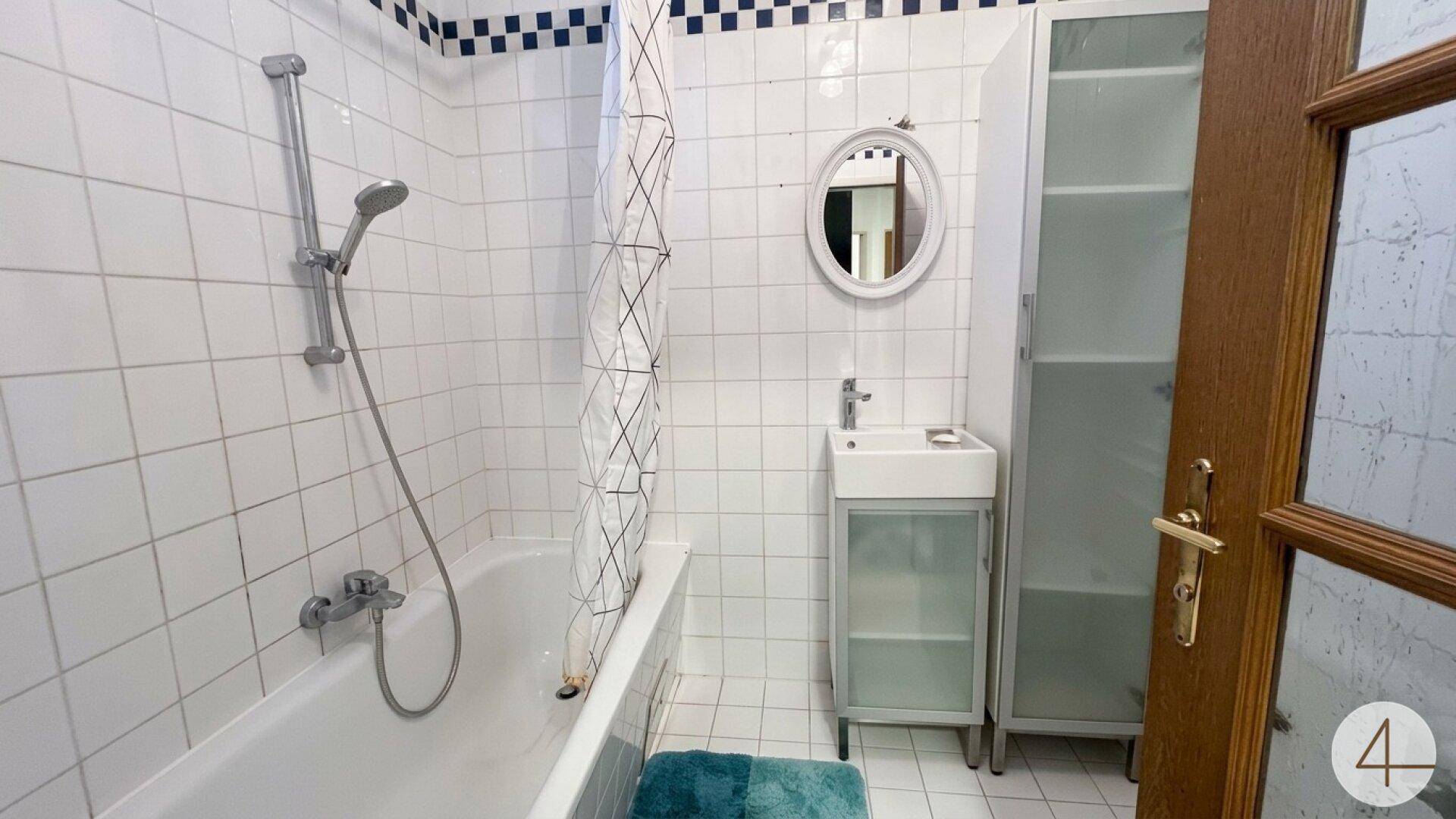 Helles Badezimmer mit weißen Fliesen, Badewanne mit Duschfunktion und kleinem Waschbecken.