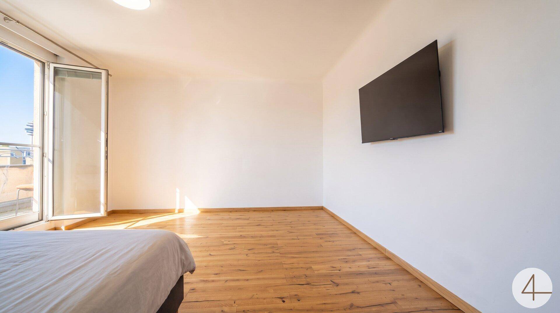 Lichtdurchflutetes Schlafzimmer mit Holzboden, Wandfernseher und Zugang zum Außenbereich.